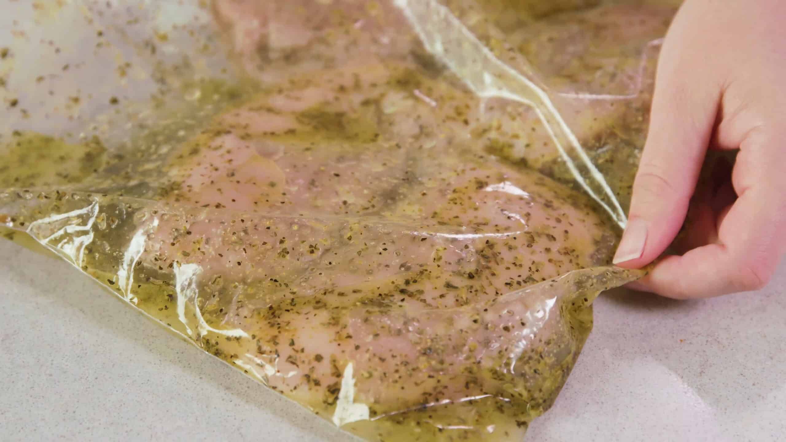 Close-up view of clear plastic seal top bag filled with lemon herb marinade and chicken breast meat to display proper coating of the meat.