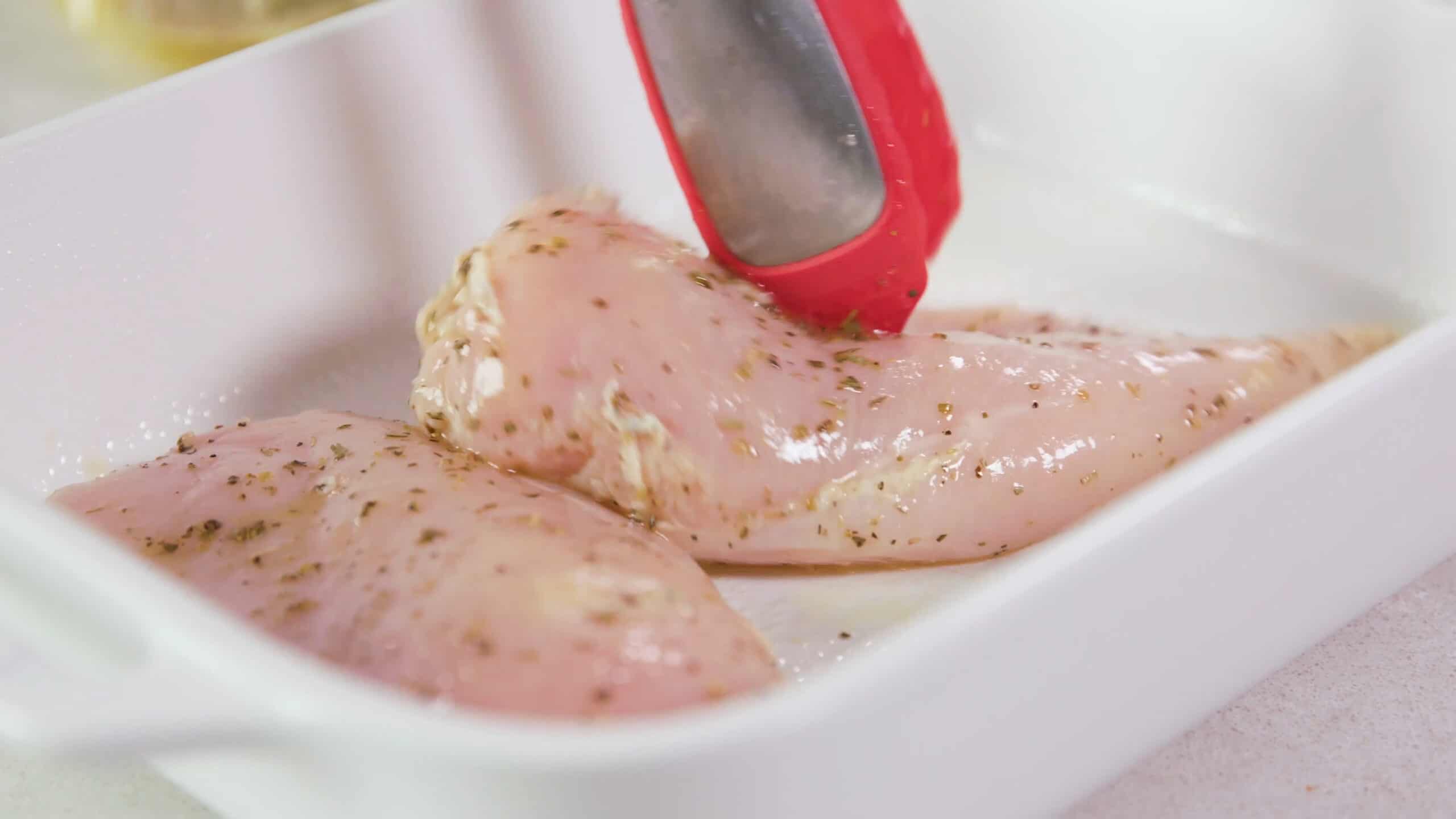 Angled view of white glass casserole dish being filled with lemon herb coated chicken breasts placed in using red plastic tongs all on a clean marble countertop.