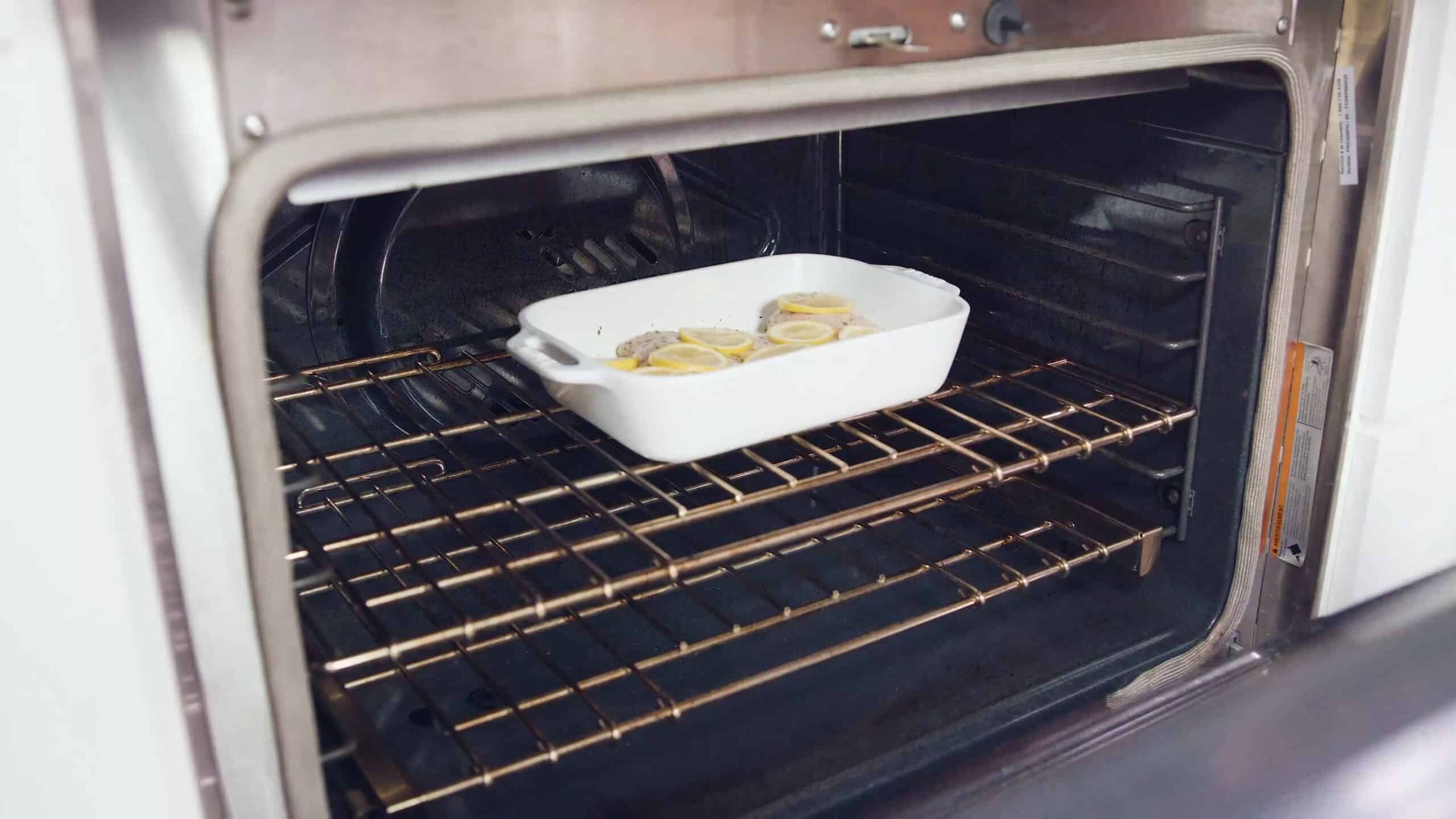 Angled view of an open oven with a white glass casserole dish filled with lemon and herb marinated chicken breasts and topped with lemon slices all on a metal rack in the middle of the oven.