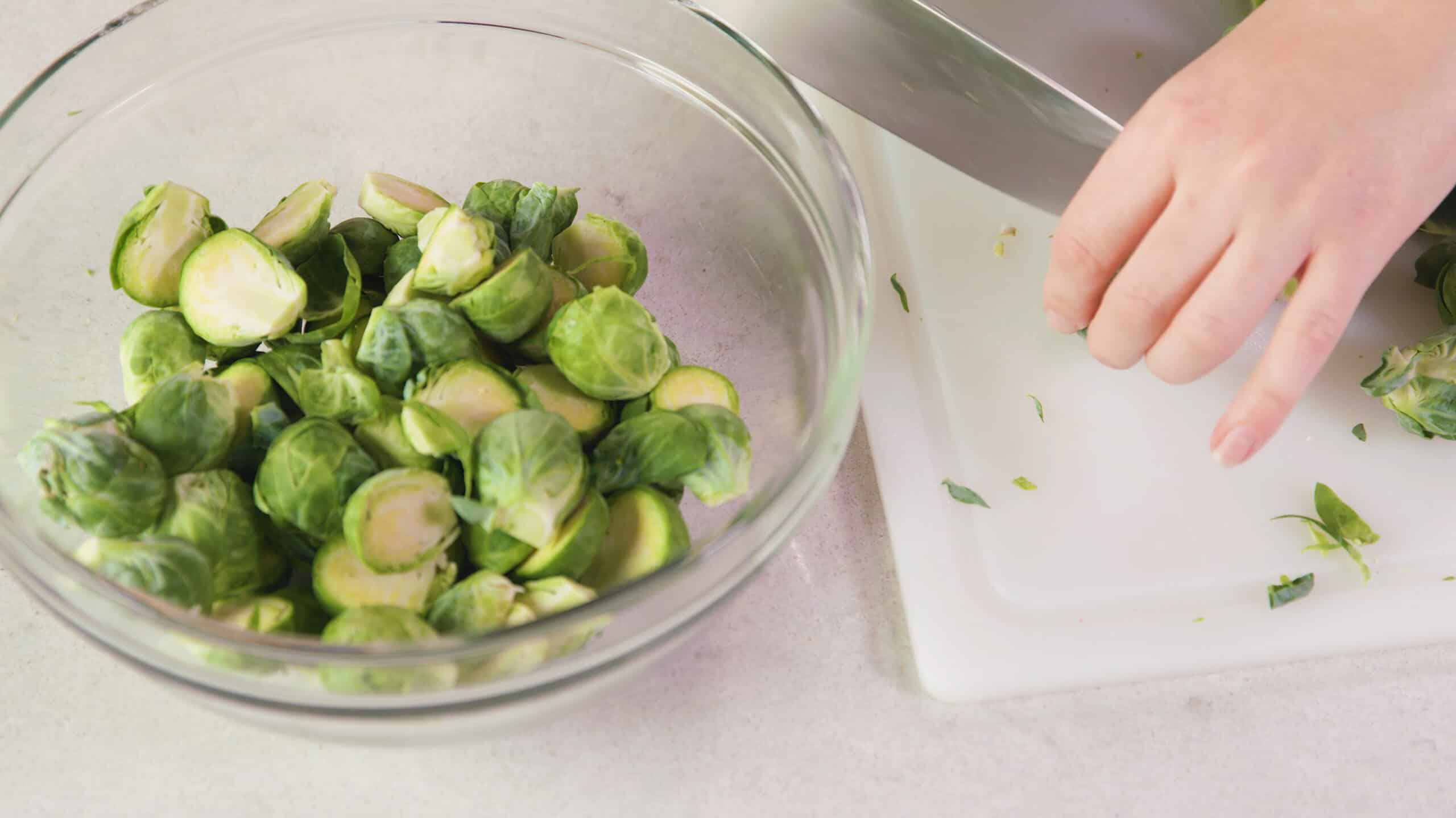 Angled view of a large clear glass mixing bowl filled with halved Brussel sprouts and a white plastic cutting board with a metal kitchen knife and a hand holding a single Brussel sprout all on a clean marble countertop.