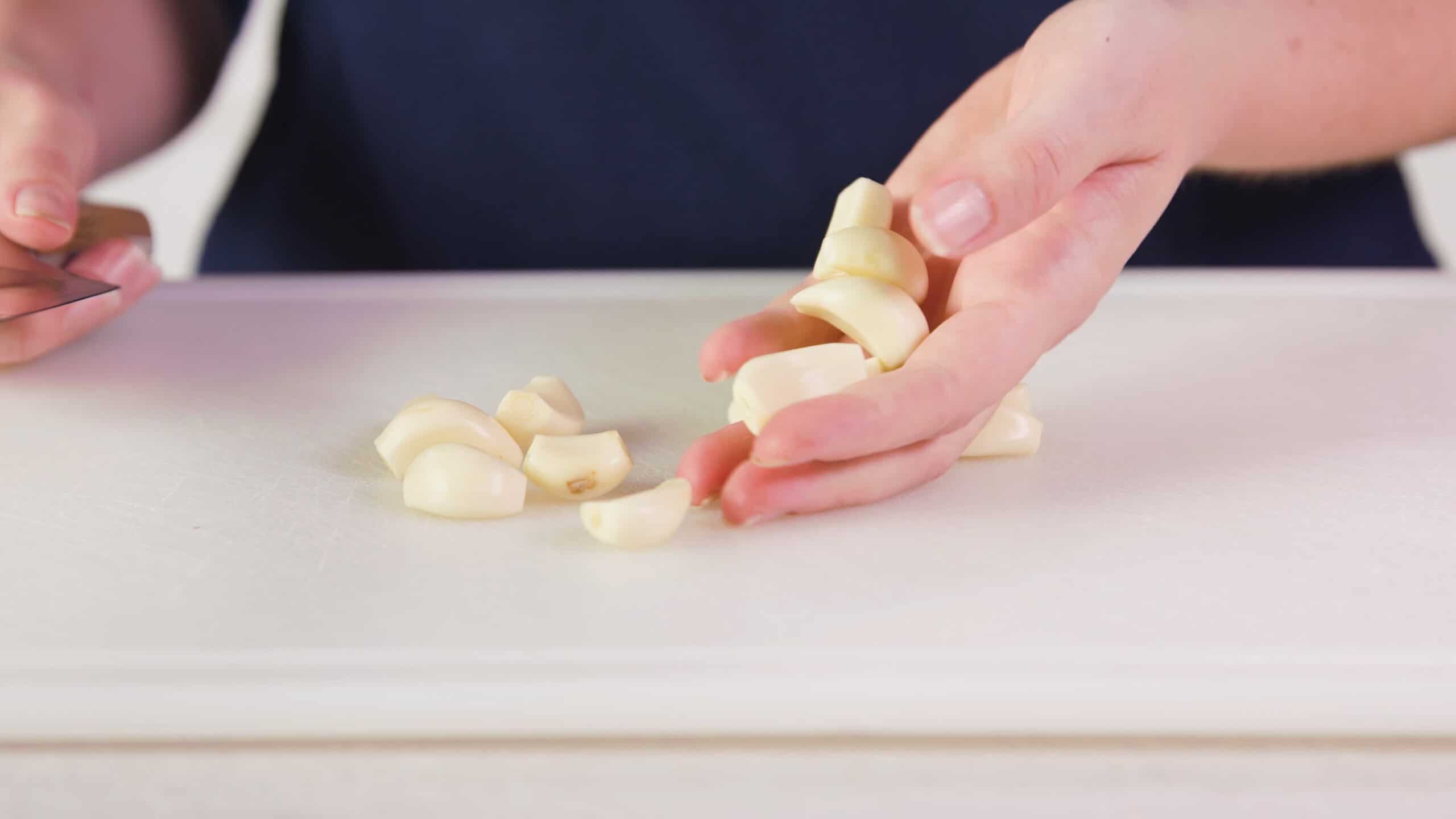 Side view of a white plastic cutting board with about one dozen garlic cloves trimmed and cleaned with a hand holding six of them all on a clean marble countertop.