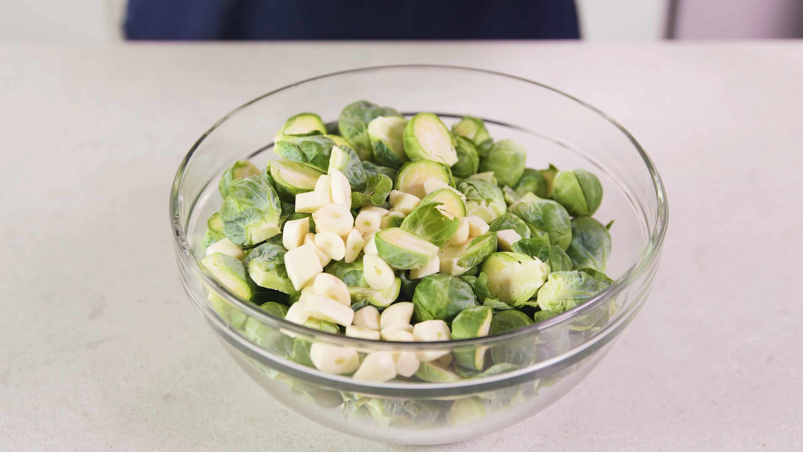 Angled view of large clear glass mixing bowl filled with sliced garlic and halved Brussel sprouts.