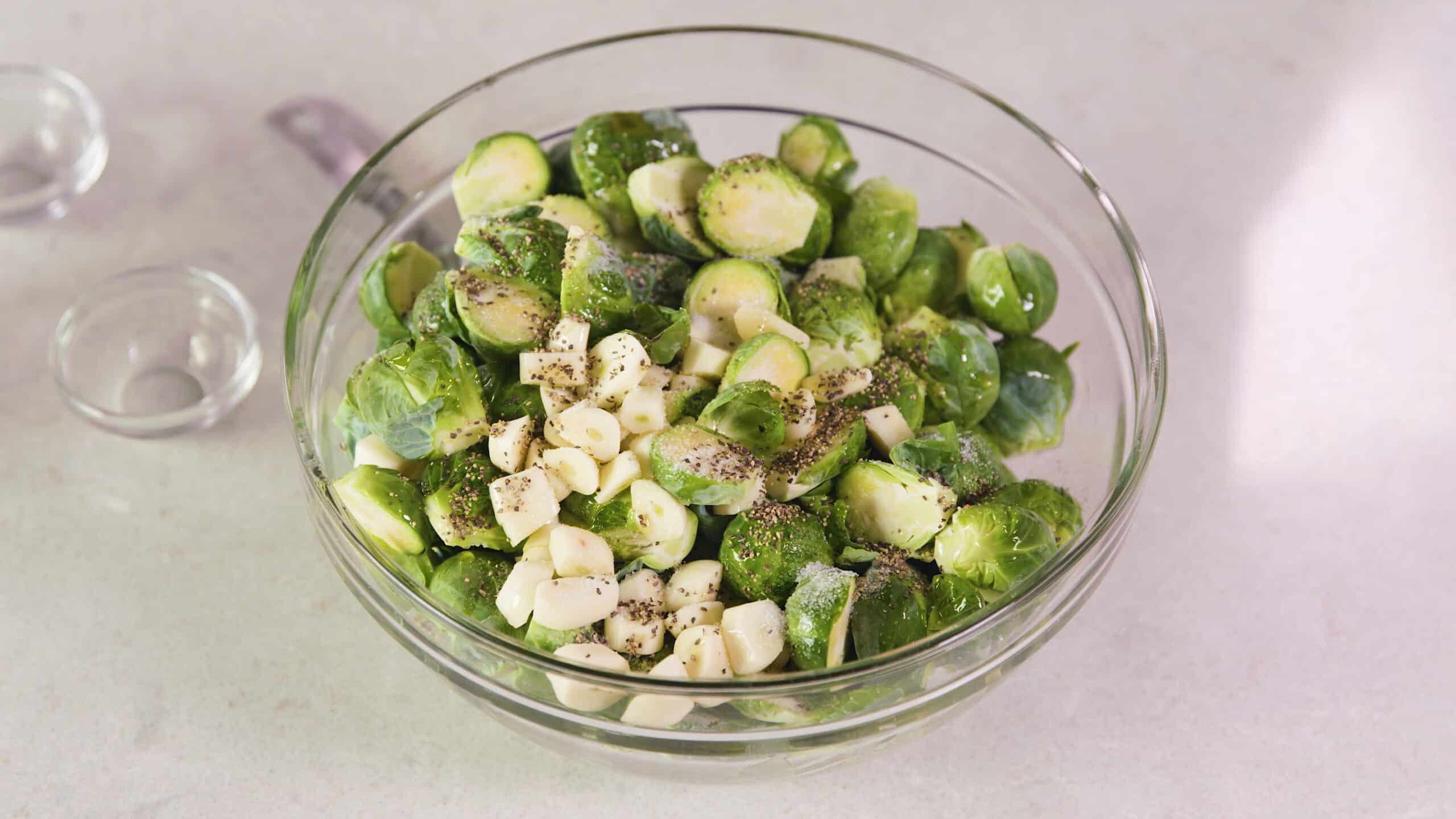 Angled view of large clear glass mixing bowl filled with sliced garlic and halved Brussel sprouts topped with olive oil, freshly ground black pepper and salt to taste.