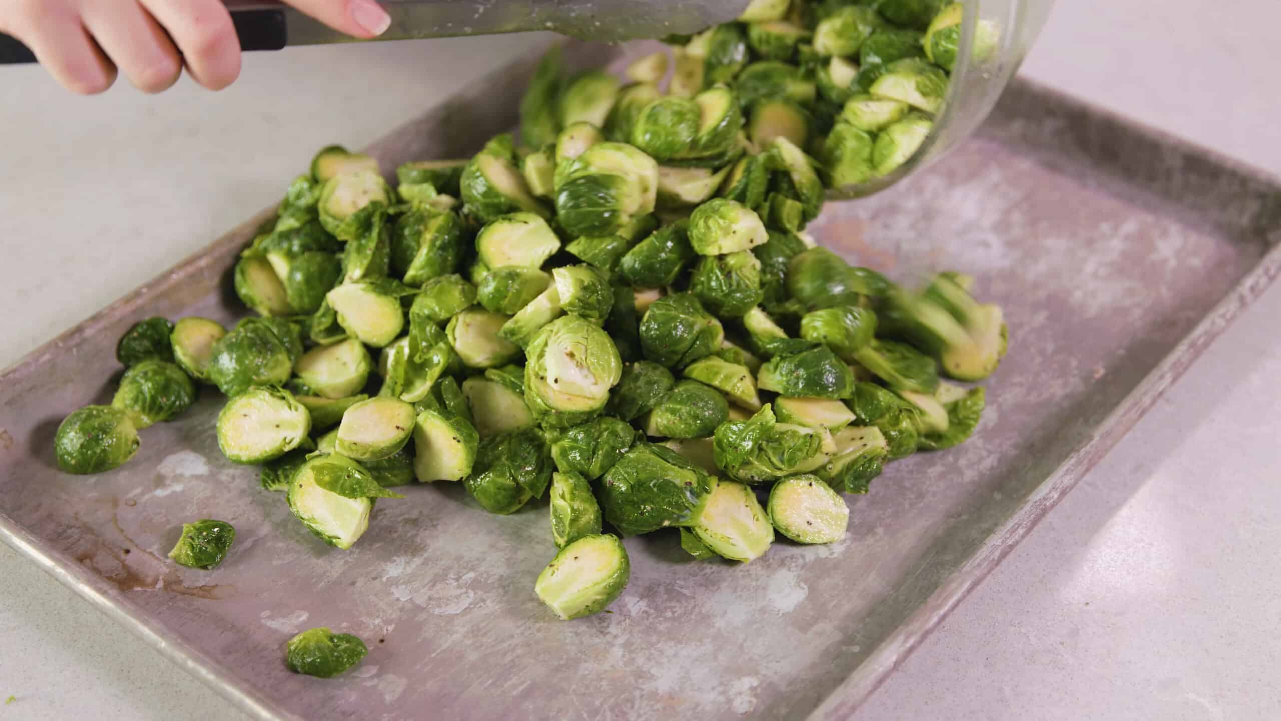 Angled view of aluminum baking sheet being filled with seasoned sliced garlic and halved Brussel sprouts being poured from a large clear glass mixing bowl.
