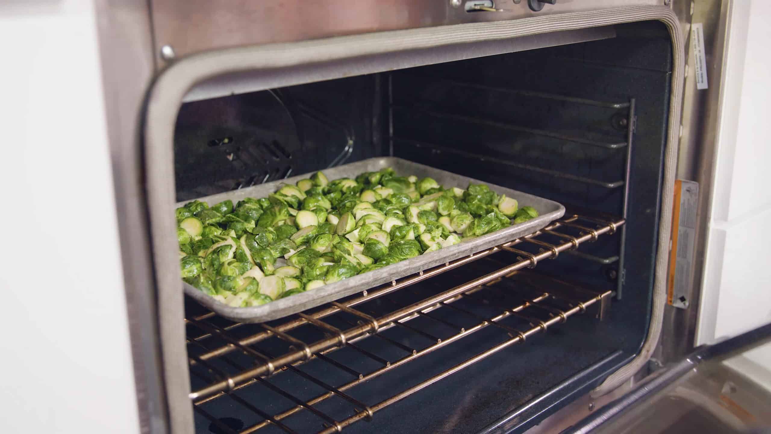 Angled view of an open oven with an aluminum baking sheet filled with seasoned sliced garlic and halved Brussel sprouts placed on a metal rack in the middle of the oven.