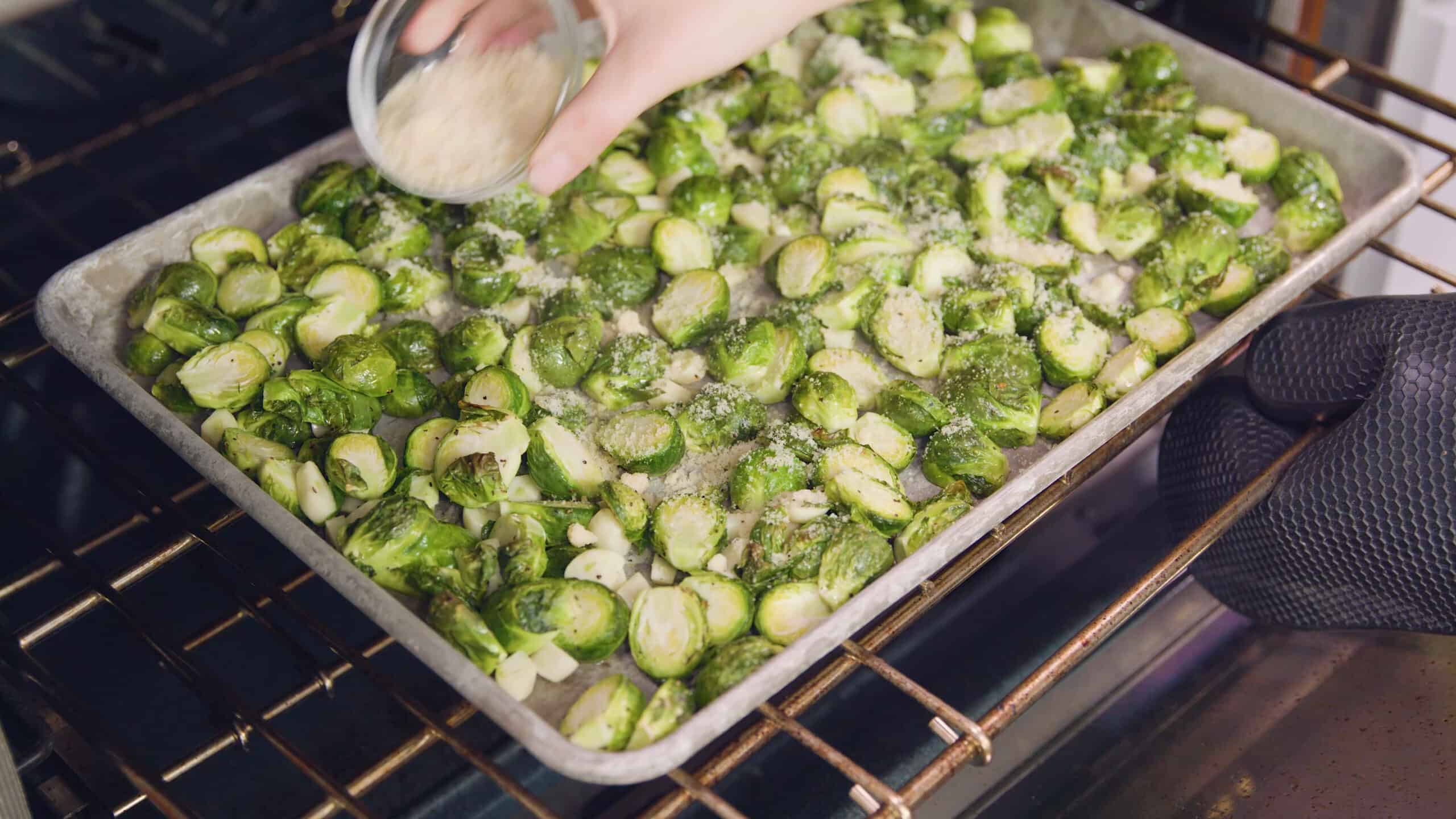 Angled view of aluminum baking sheet filled with seasoned sliced garlic and halved Brussel sprouts and grated parmesan cheese poured from a small clear glass mixing bowl on top.