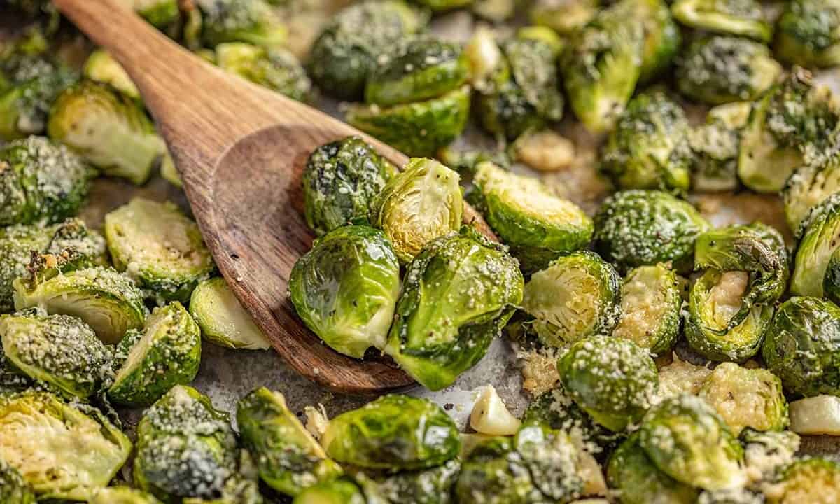 A wooden spoon scoops up Garlic Butter Roasted Brussel Sprouts from a sheet pan.