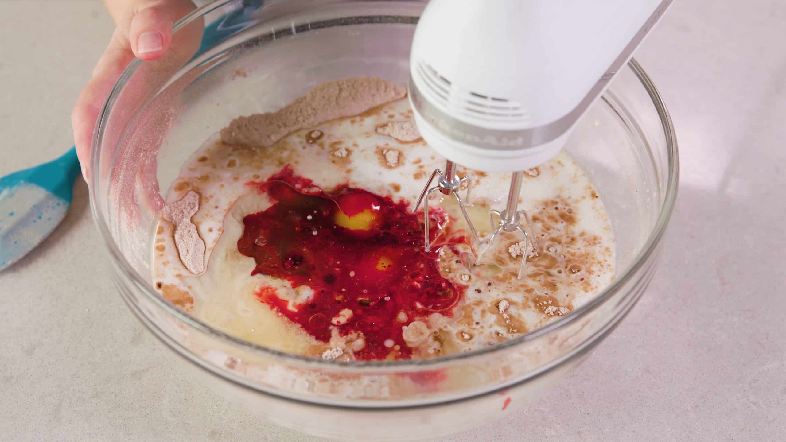 Angled view of clear glass mixing bowl filled with wet and dry ingredients including red food coloring with wire beaters attached to a hand mixer ready to combine.