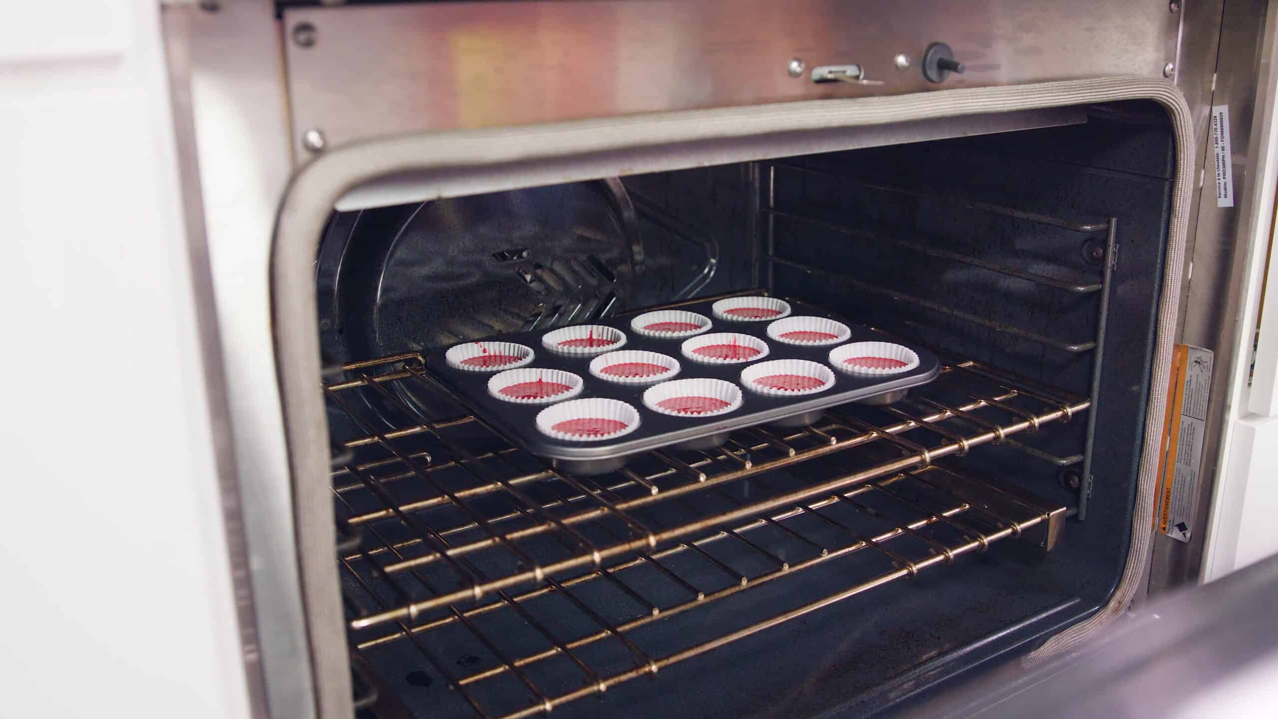 Angled view of an open oven with metal muffin tin with one dozen paper liners filled with red velvet cake batter placed on a metal rack in the middle of the oven.