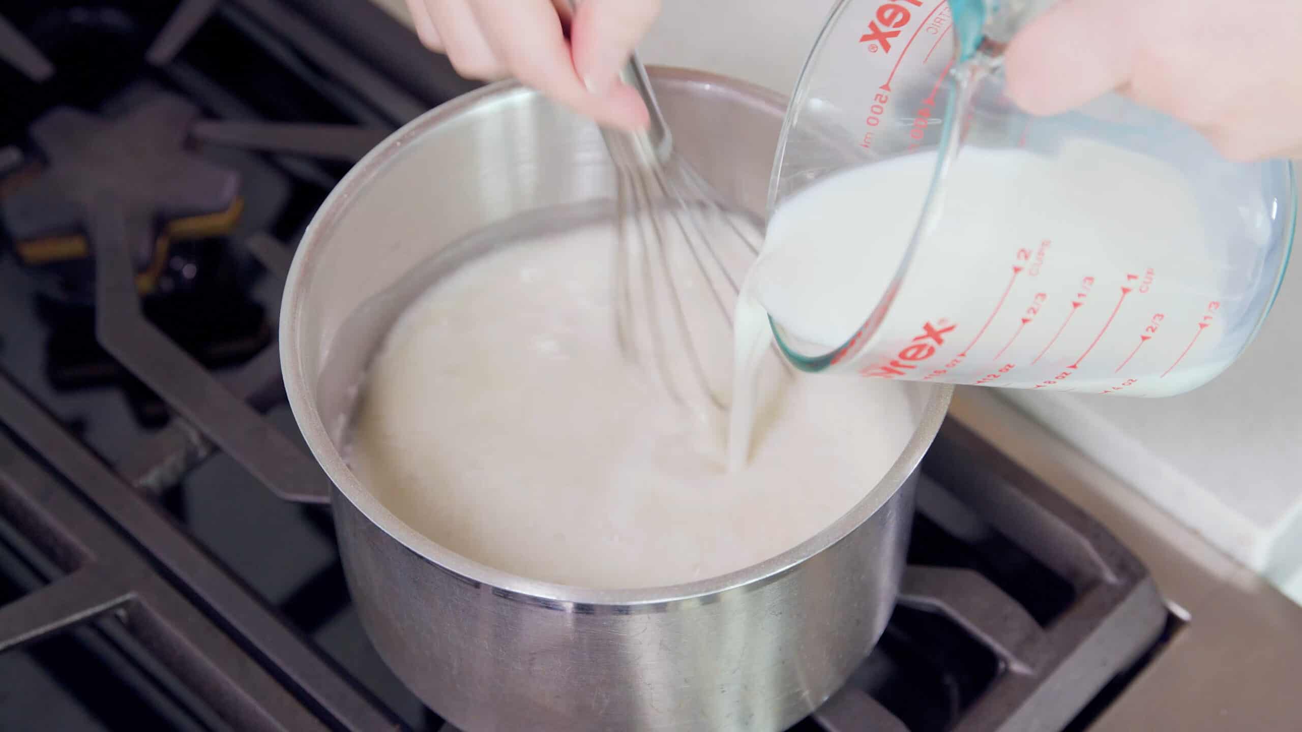 Angled view of metal pot filled with various wet ingredients for Ermine Icing with whole milk being poured from a clear glass measuring cup and a wire whisk stirring the ingredients to combine.