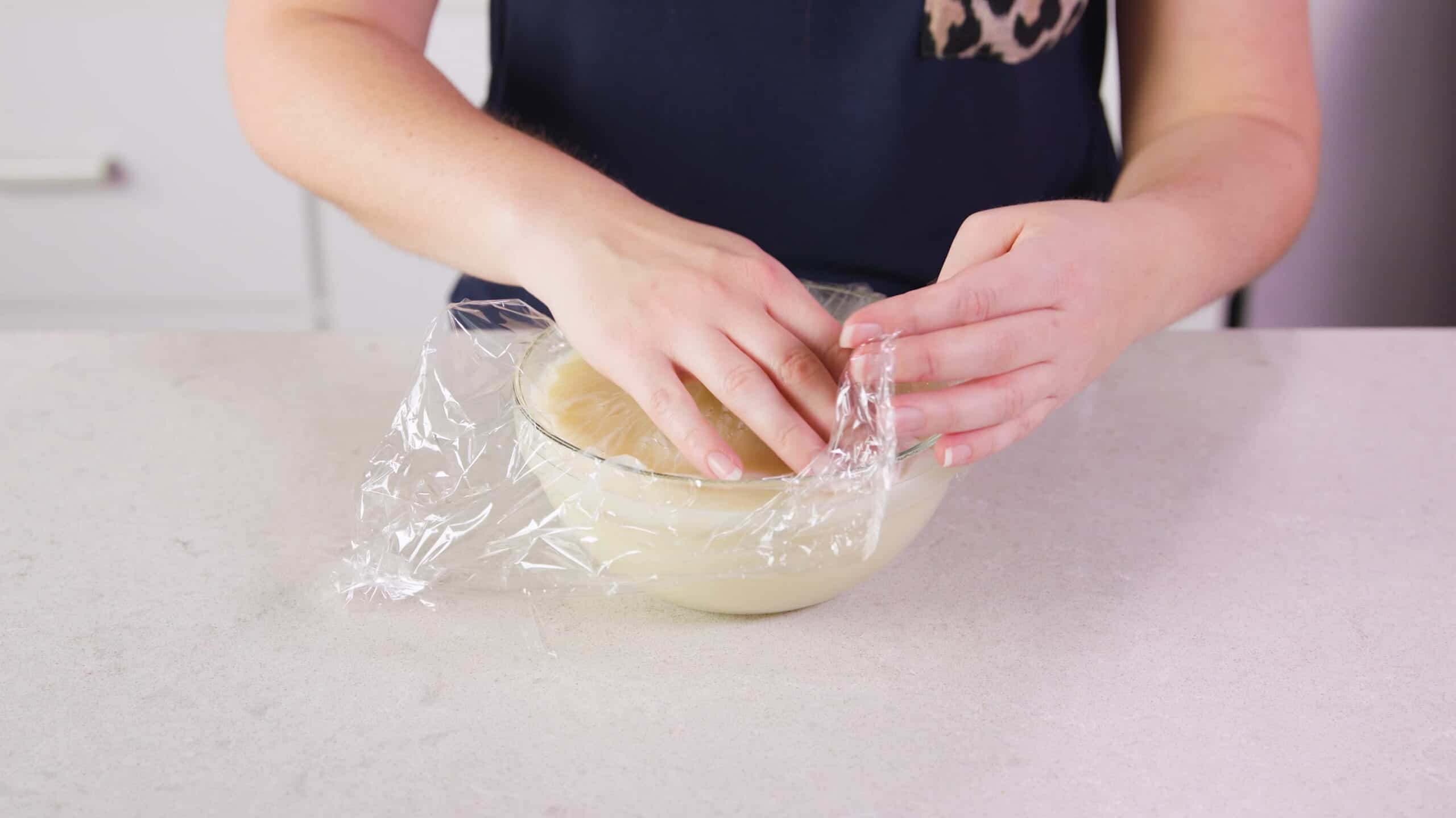 Angled view of clear glass mixing bowl filled with Ermine Icing and topped with clear plastic wrap to keep the top from stiffening while set aside.