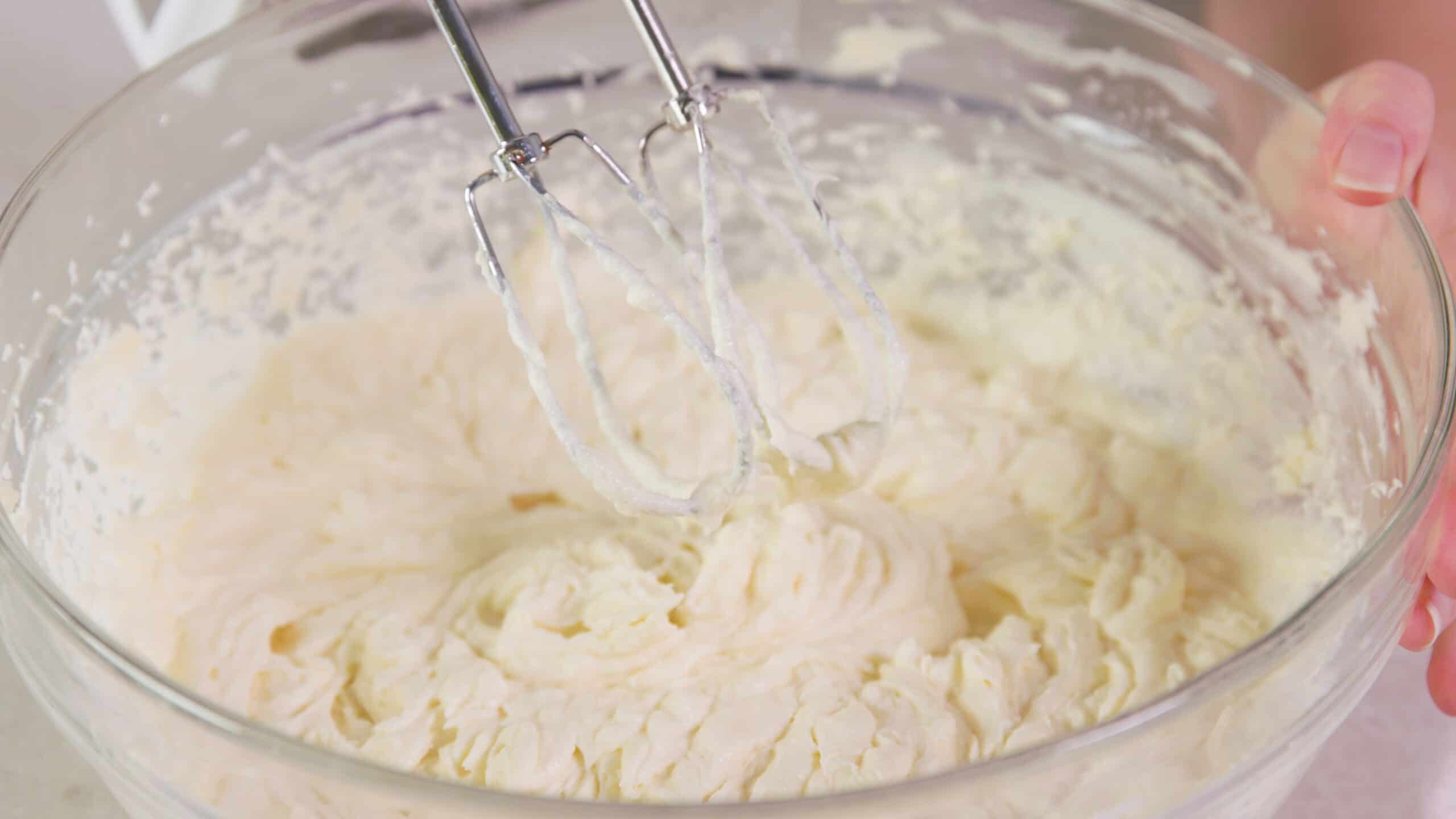 Close-up view of a large clear glass mixing bowl filled with whipped butter and milk mixture to complete Ermine Icing in preparation for topping the red velvet cupcakes.