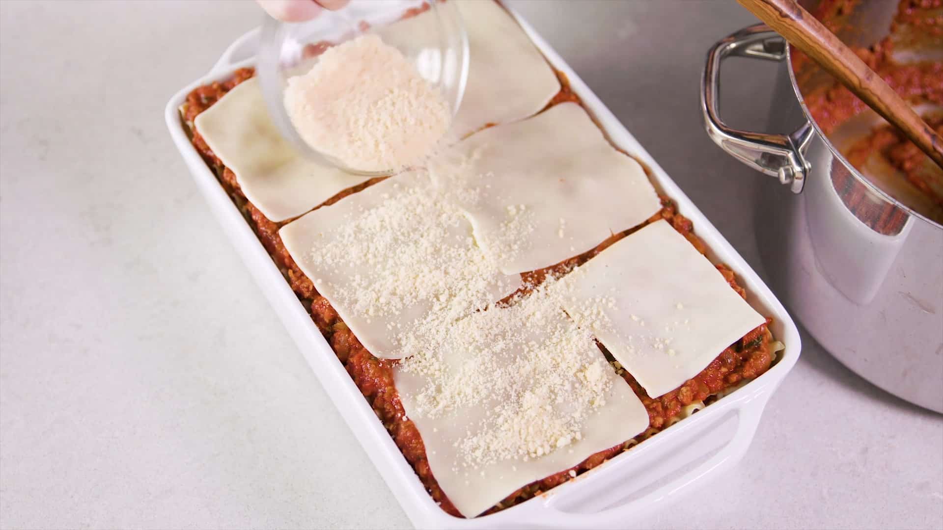 Overhead view of a high sided white glass casserole dish filled with multiple layers of lasagna and the newest layer being added to the top layer of mozzarella cheese slices with grated parmesan cheese sprinkled from a clear glass mixing bowl.
