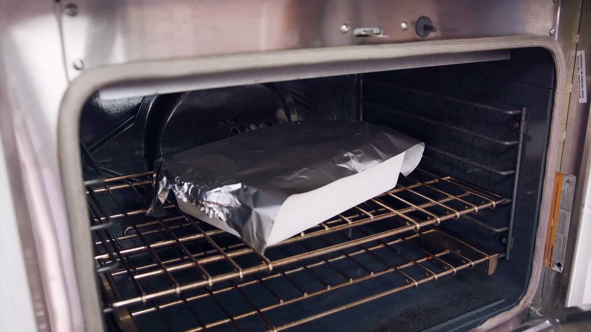 Angled view of an open oven with a high sided white glass casserole dish topped with a long sheet of aluminum foil shiny side facing down all on a metal rack in the middle of the oven.