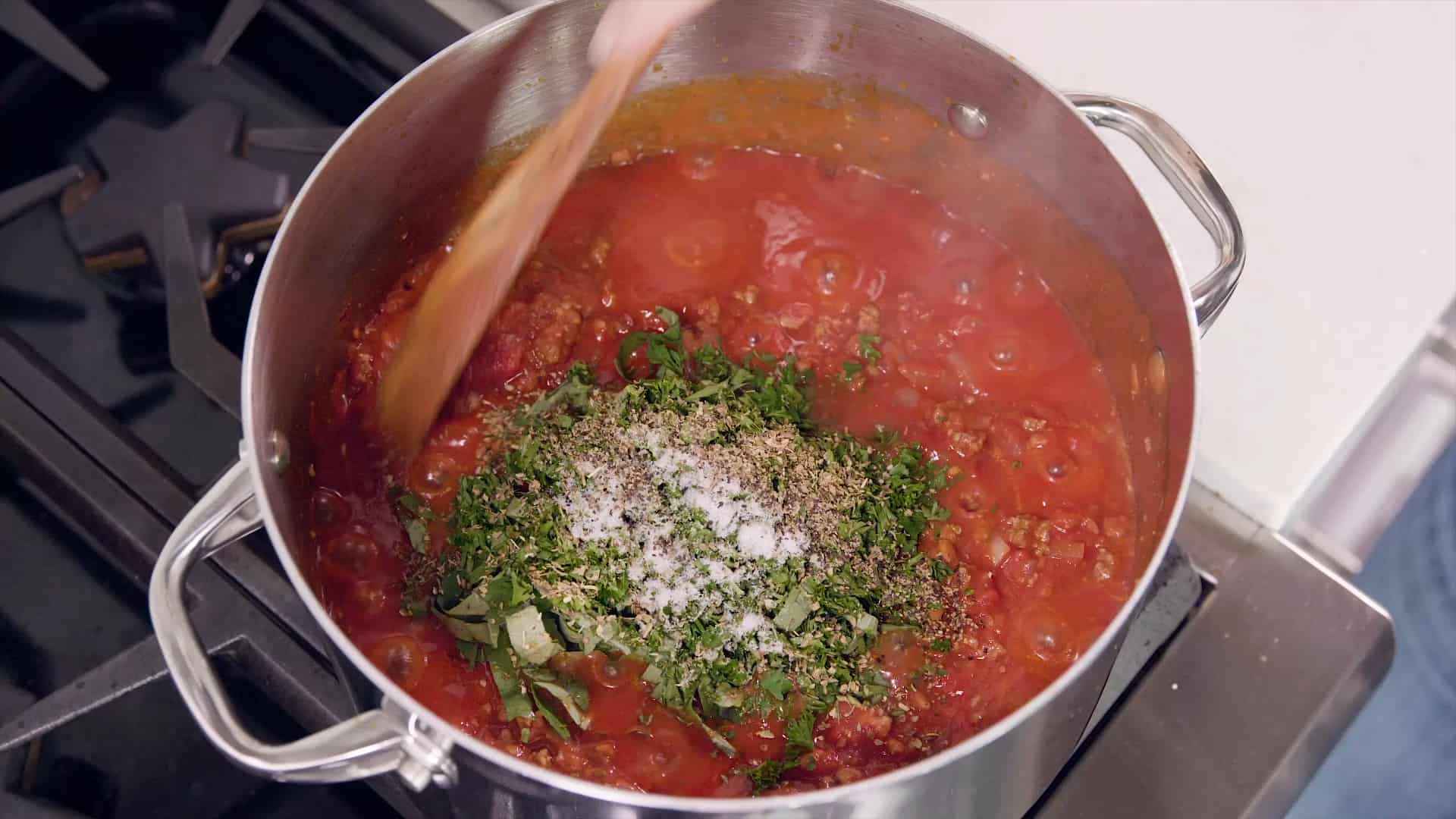 Overhead view of metal saucepan filled with red sauce and fresh chopped oregano, chopped basil and other dried spices and a wooden spoon stirring to combine all stirring on a stovetop burner.