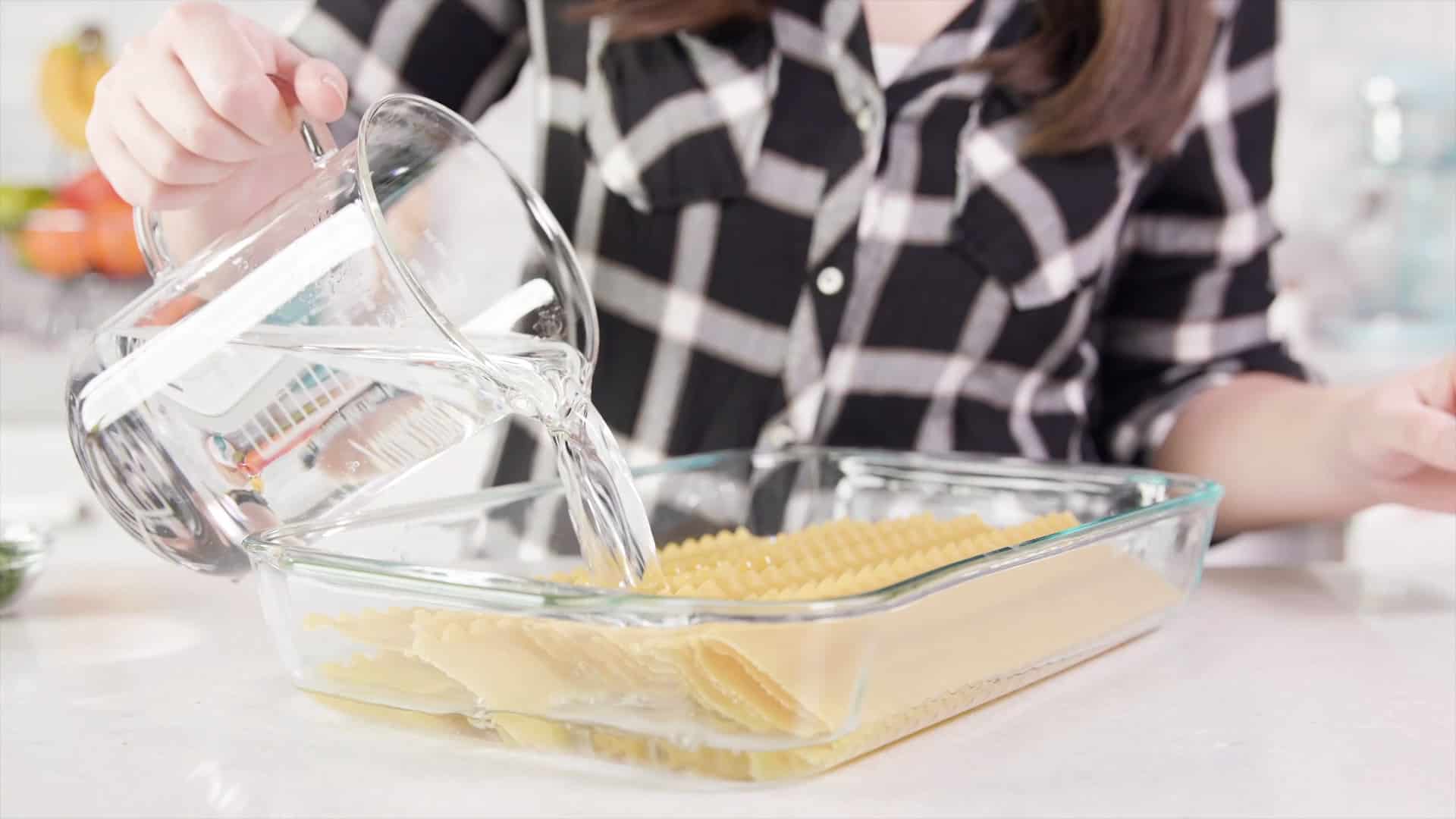 Angled view of a clear glass casserole dish filled with dry lasagna noodles being filled with water poured from a large clear glass measuring cup.