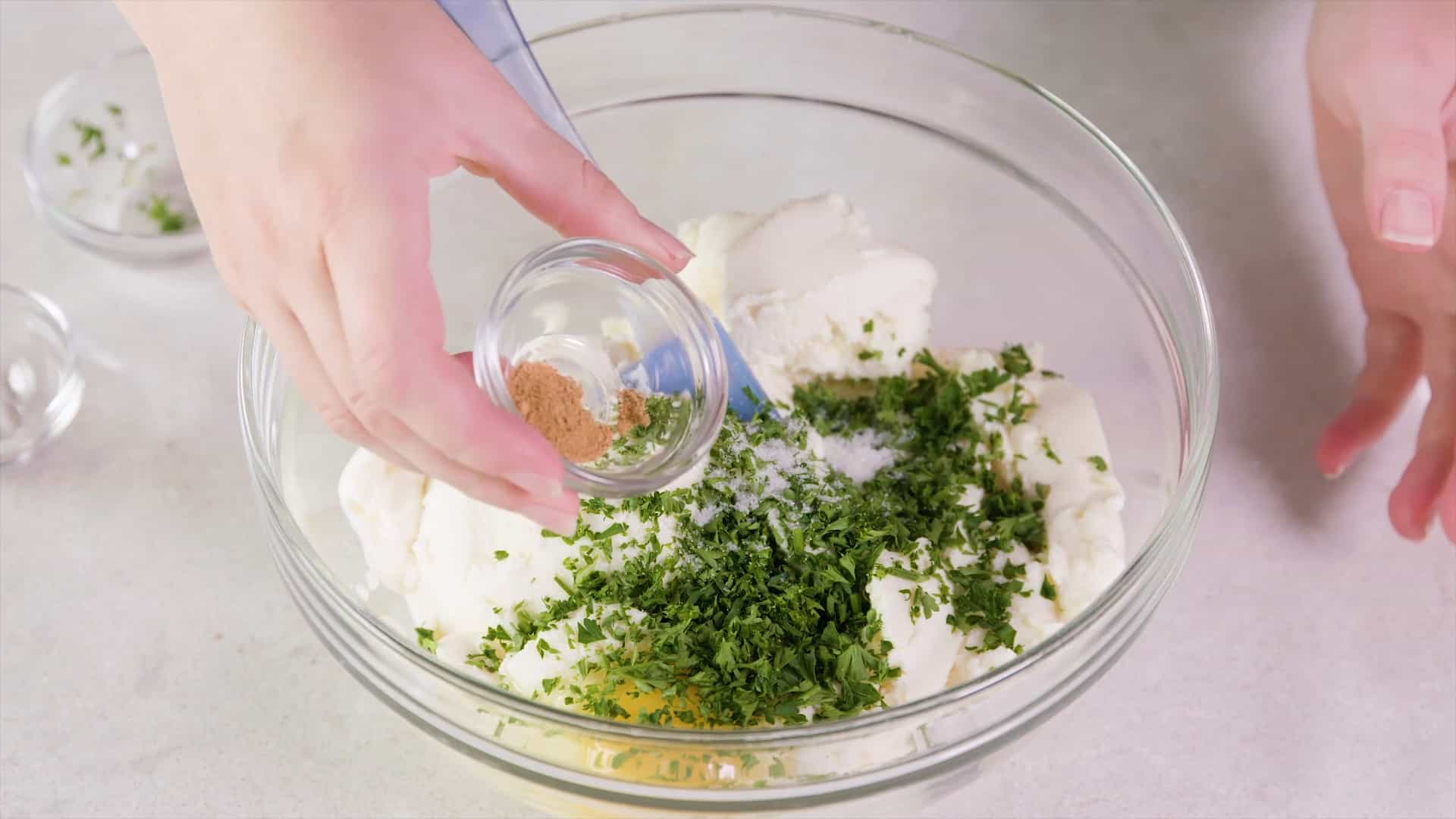 Overhead view of a large clear glass mixing bowl filled with ricotta cheese mixture and topped with an egg, chopped parsley and salt to taste, with a small clear glass bowl with nutmeg ready to pour with a plastic spatula at the ready to combine.
