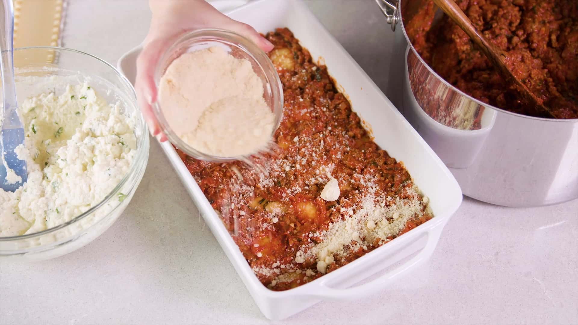 Overhead view of a high sided white glass casserole dish filled with multiple layers of lasagna and the newest layer being added to the meat sauce layer with grated parmesan cheese sprinkled from a medium sized clear glass mixing bowl.