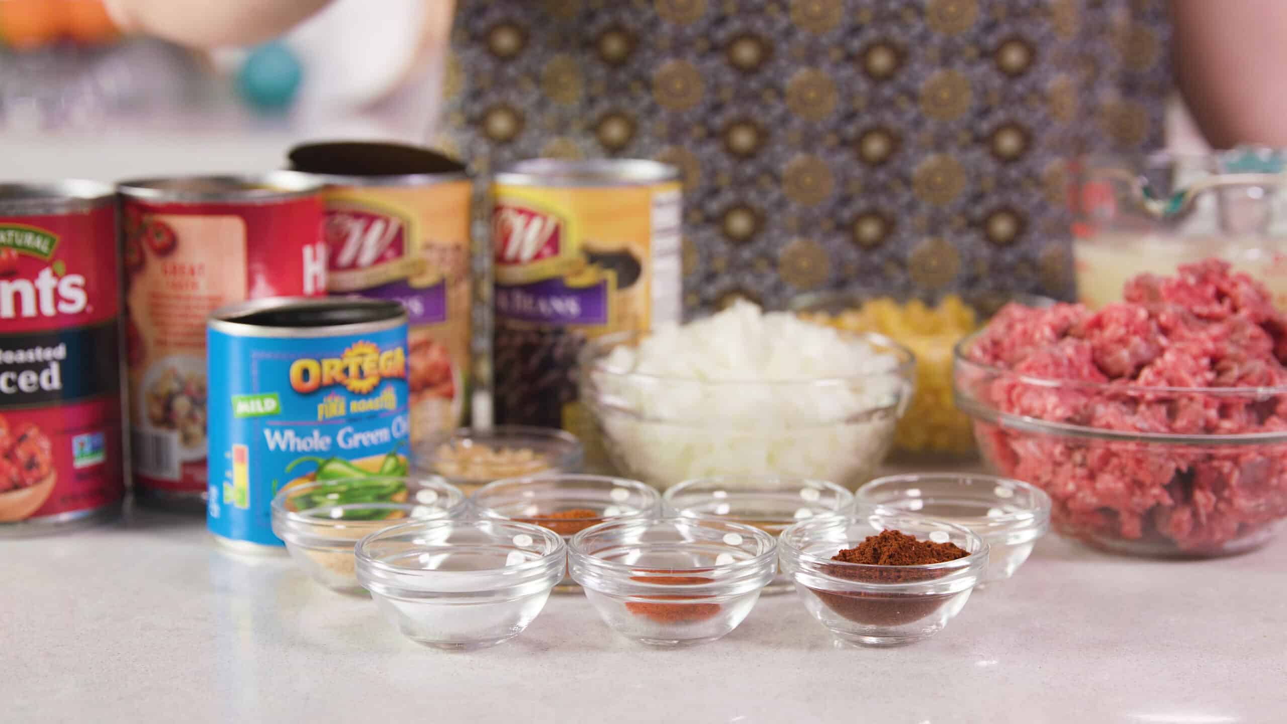 Side view of all the ingredients needed to complete the taco soup recipe. Some ingredients are spices in the foreground separated into clear glass bowls with some canned ingredients in the background and beef, onion, and corn in medium sized clear glass mixing bowls.