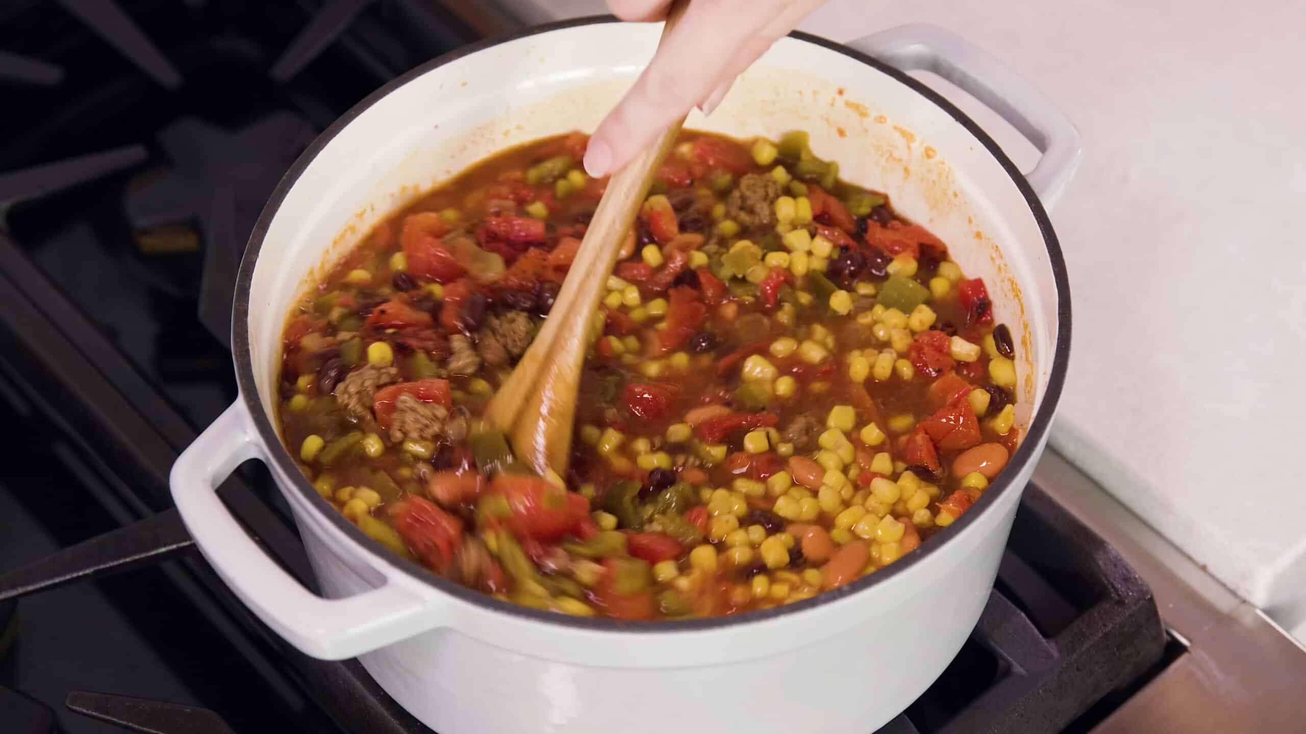 Angled view of a white enamel coated cast iron pot filled with taco soup ingredients and simmering on a stovetop burner being stirred with a wooden spoon.