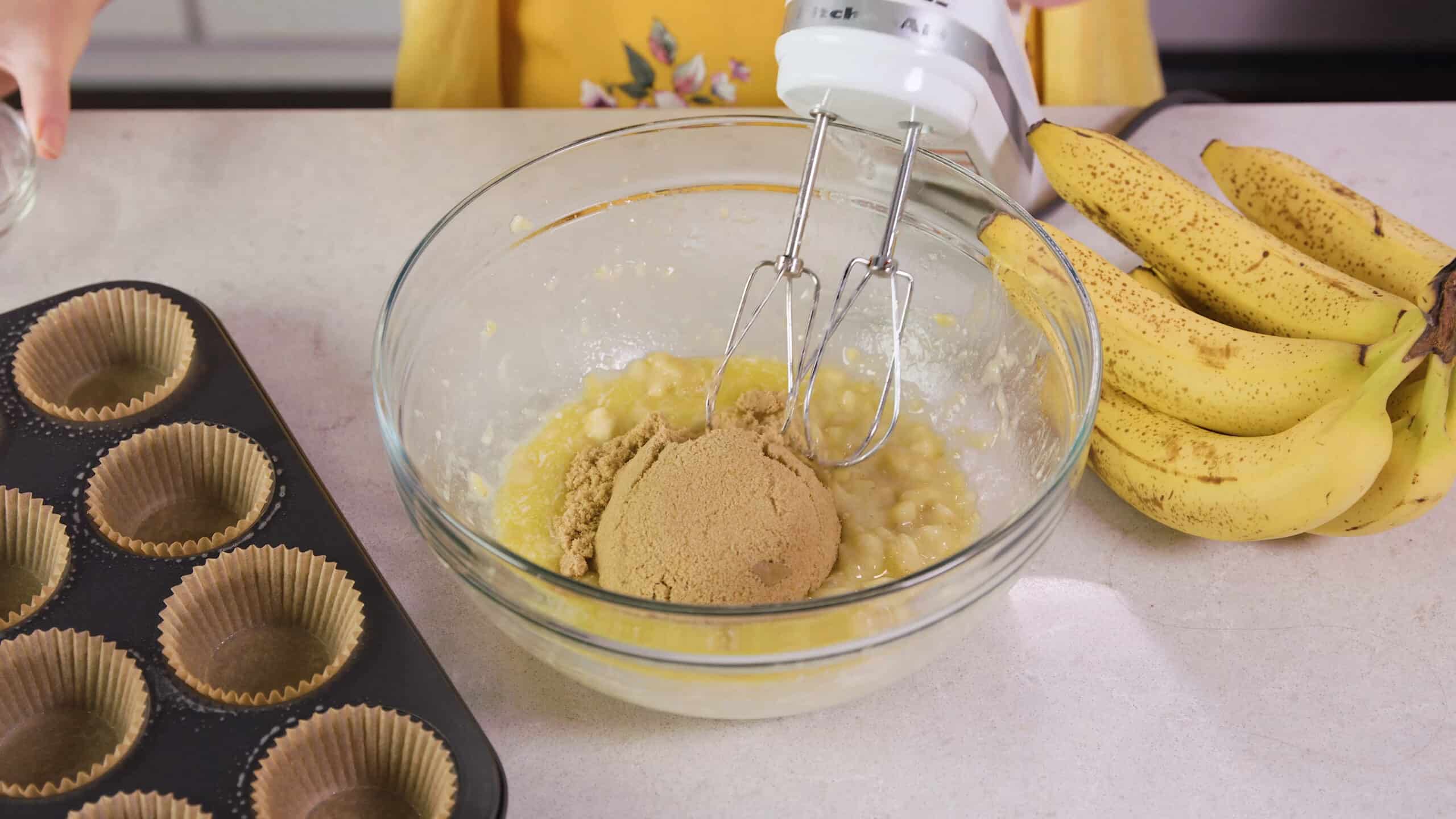 Angled view of a large clear glass mixing bowl filled with mashed bananas and brown sugar with wire beaters connected to a hand mixer ready to combine.