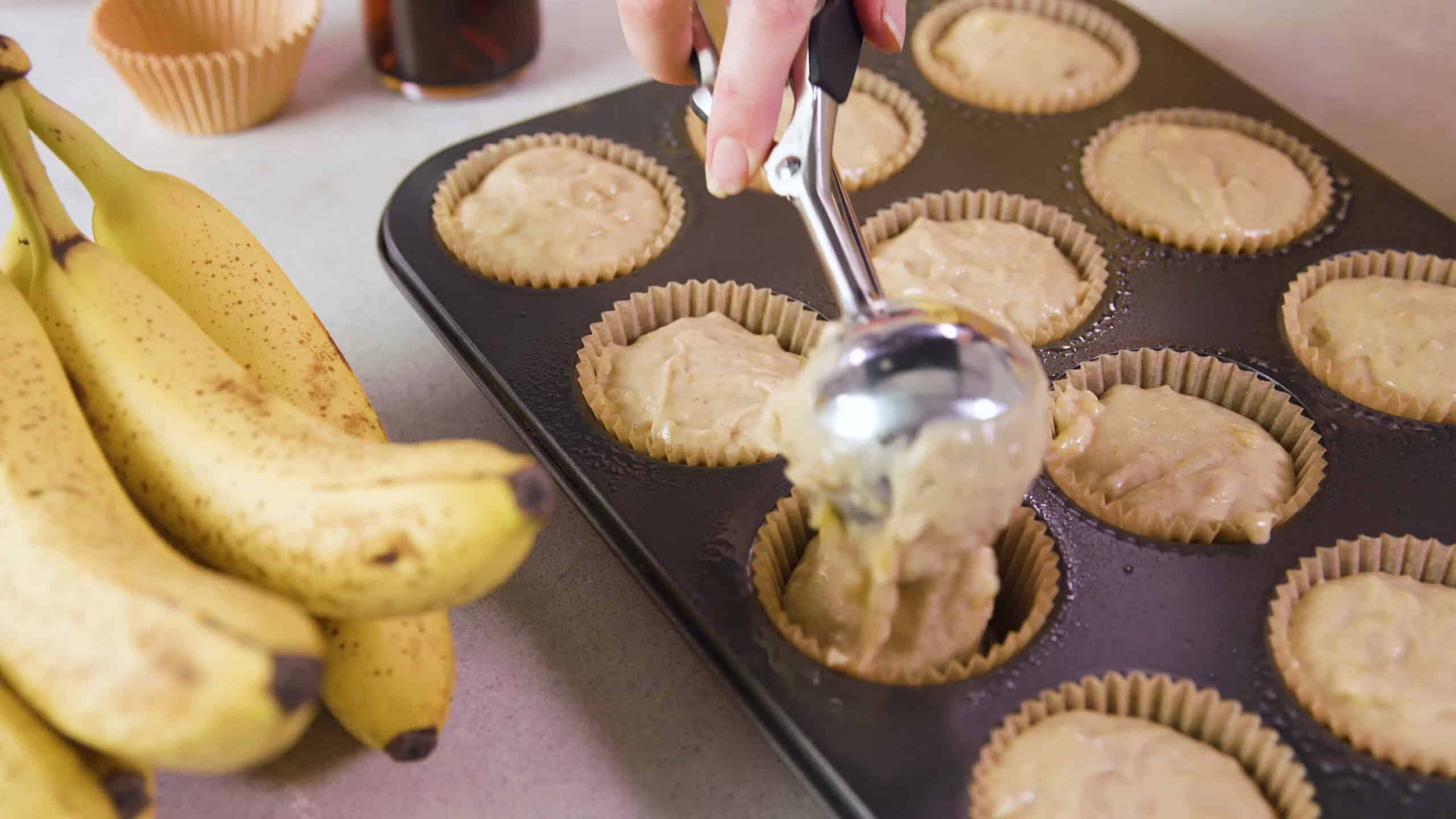 Angled view of metal muffin tin filled with paper liners and greased with oil spray and being filled with banana muffin batter scooped in with a metal scoop.