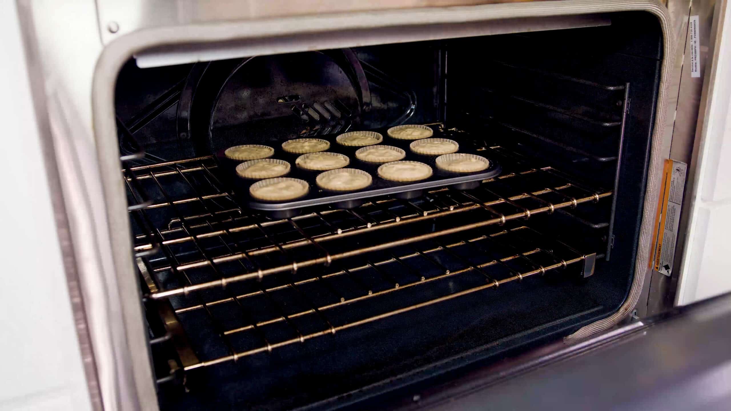 Angled view of an open oven with a metal muffin tin lined with one dozen paper liners all filled with banana muffin batter placed on a metal rack in the middle of the oven.