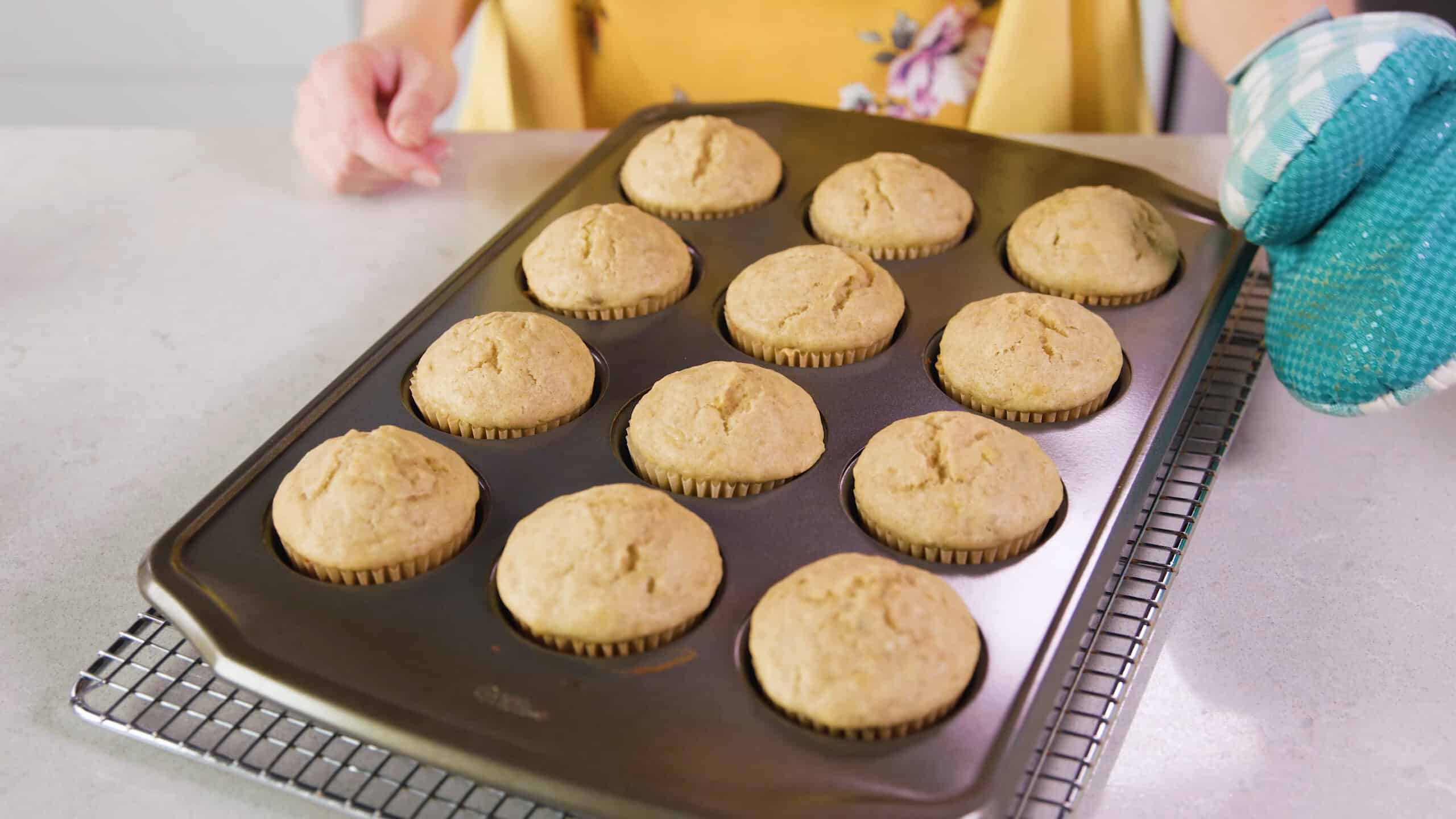 Angled view of metal muffin tin filled with one dozen cooked banana muffins with paper liners setting on a cooling rack all on a marble countertop.
