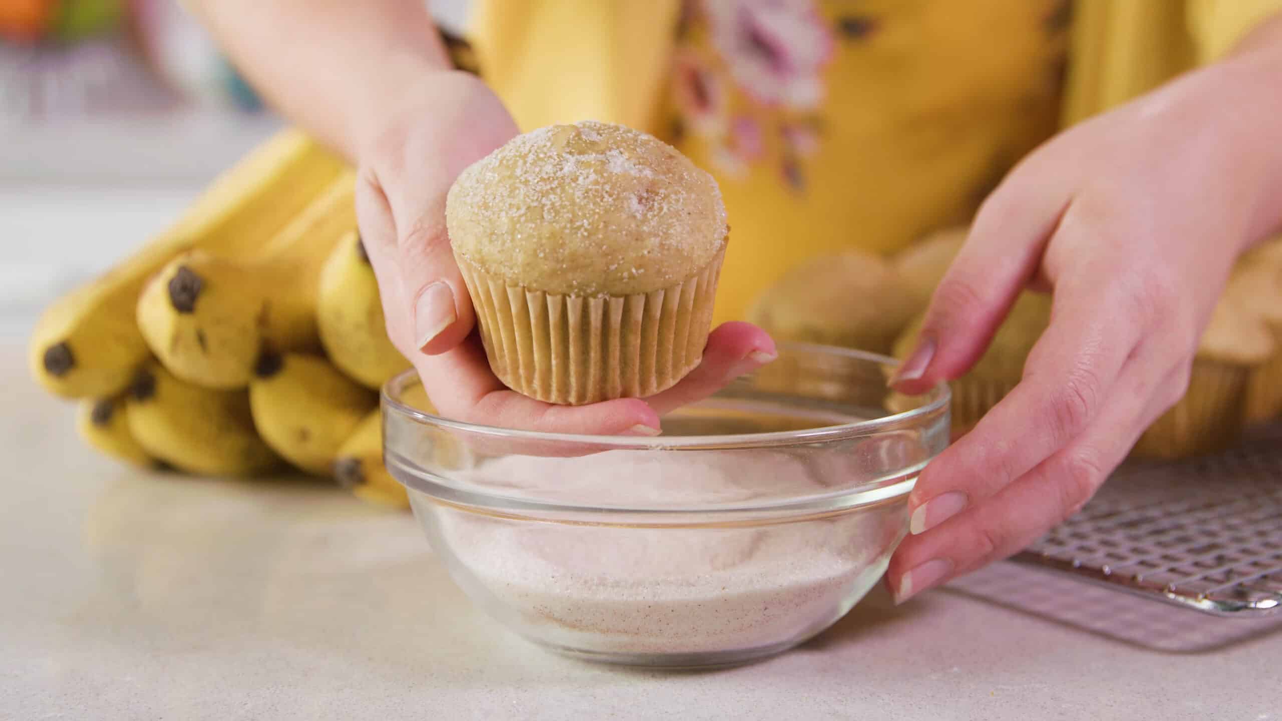 Side view of a clear glass mixing bowl filled with cinnamon and sugar with a single banana muffin held above the bowl after dipping the sugar cinnamon mixture by hand.
