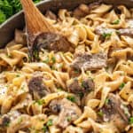 Beef stroganoff with sliced mushrooms on egg noodles stirred with a wooden spoon, parsley in background.