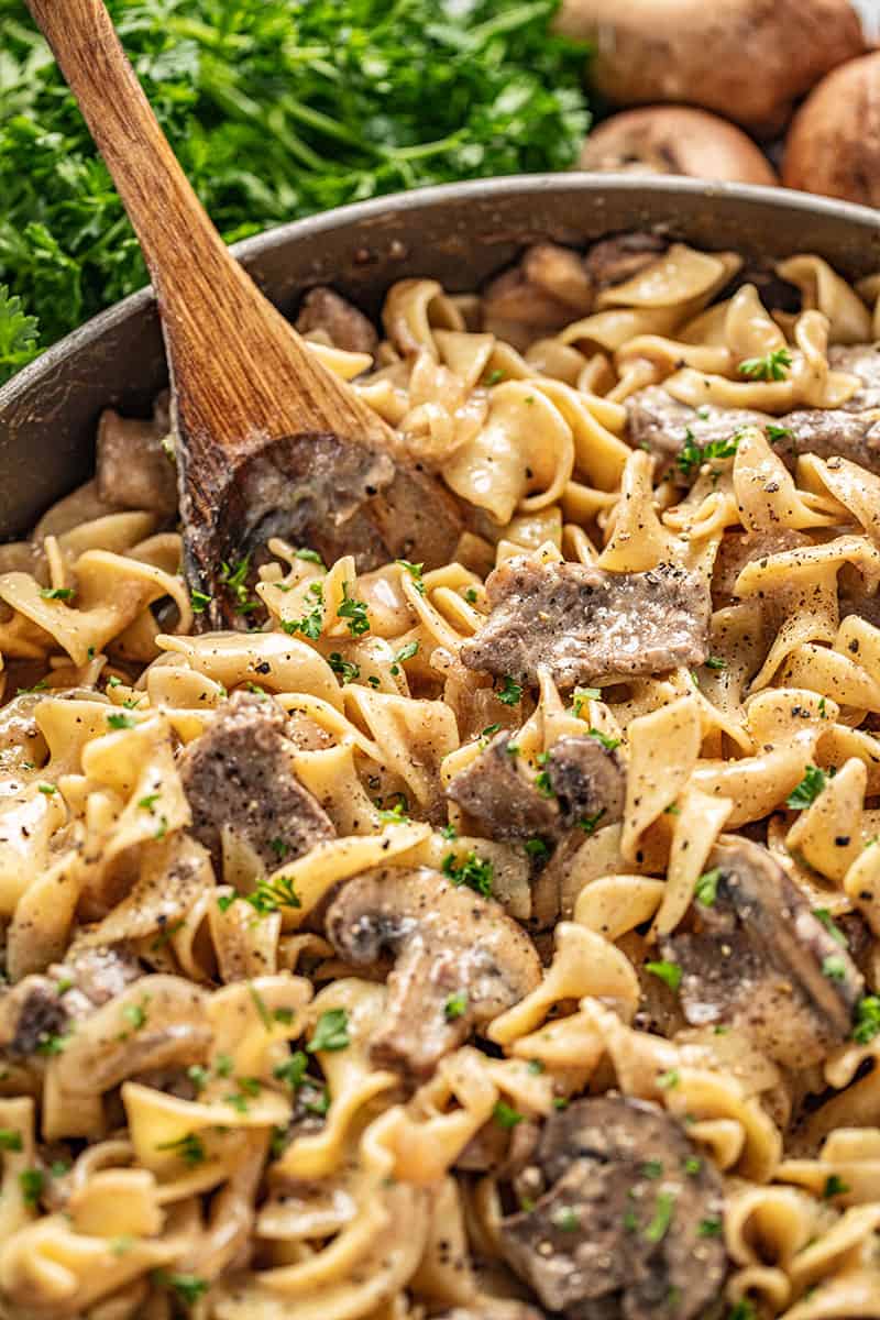 Beef stroganoff with sliced mushrooms on egg noodles stirred with a wooden spoon, parsley in background