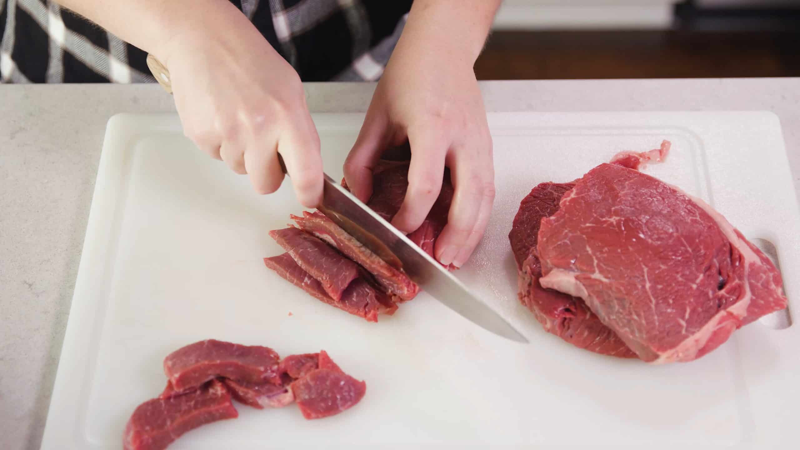 Overhead view of raw beef sirloin being cut with a kitchen knife into strips on a clean cutting board.