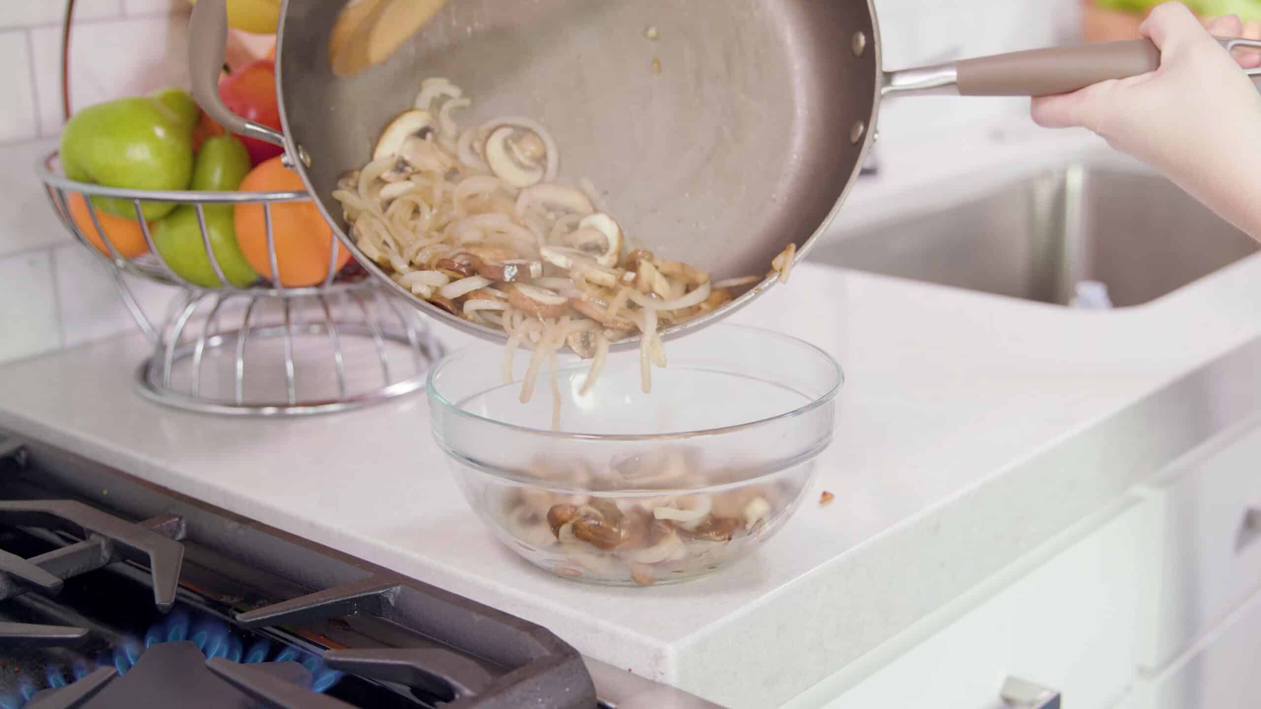 Angled view of a large glass mixing bowl on a marble countertop with sautéed onions and mushroom pouring from a metal saucepan that has been removed from a stovetop burner seen from the side.