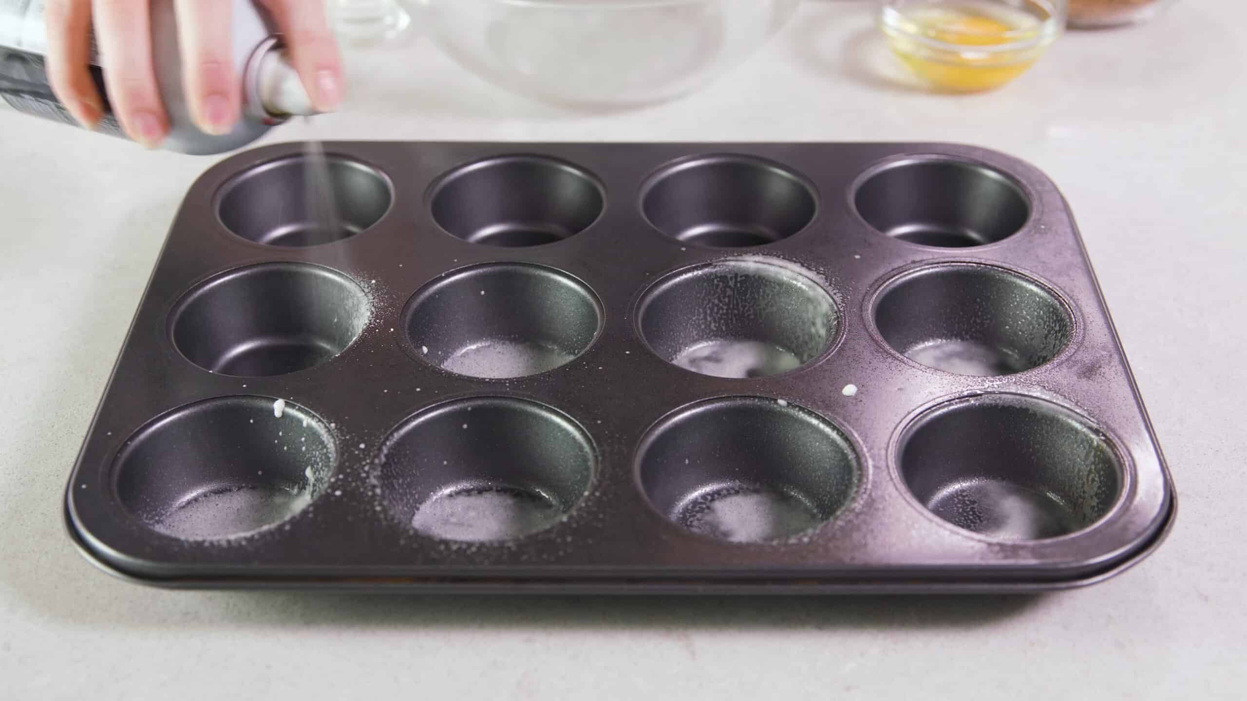 Angled view of metal muffin tin being sprayed with oil.