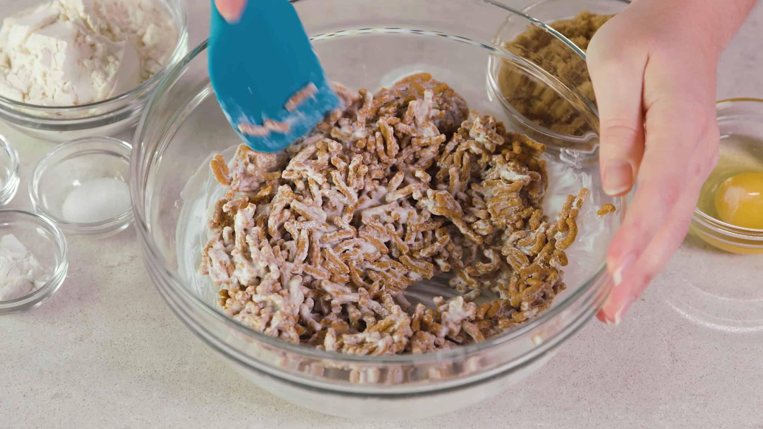 Close-up view of a large clear glass mixing bowl filled with 100% bran cereal mixed with buttermilk with a plastic spatula.