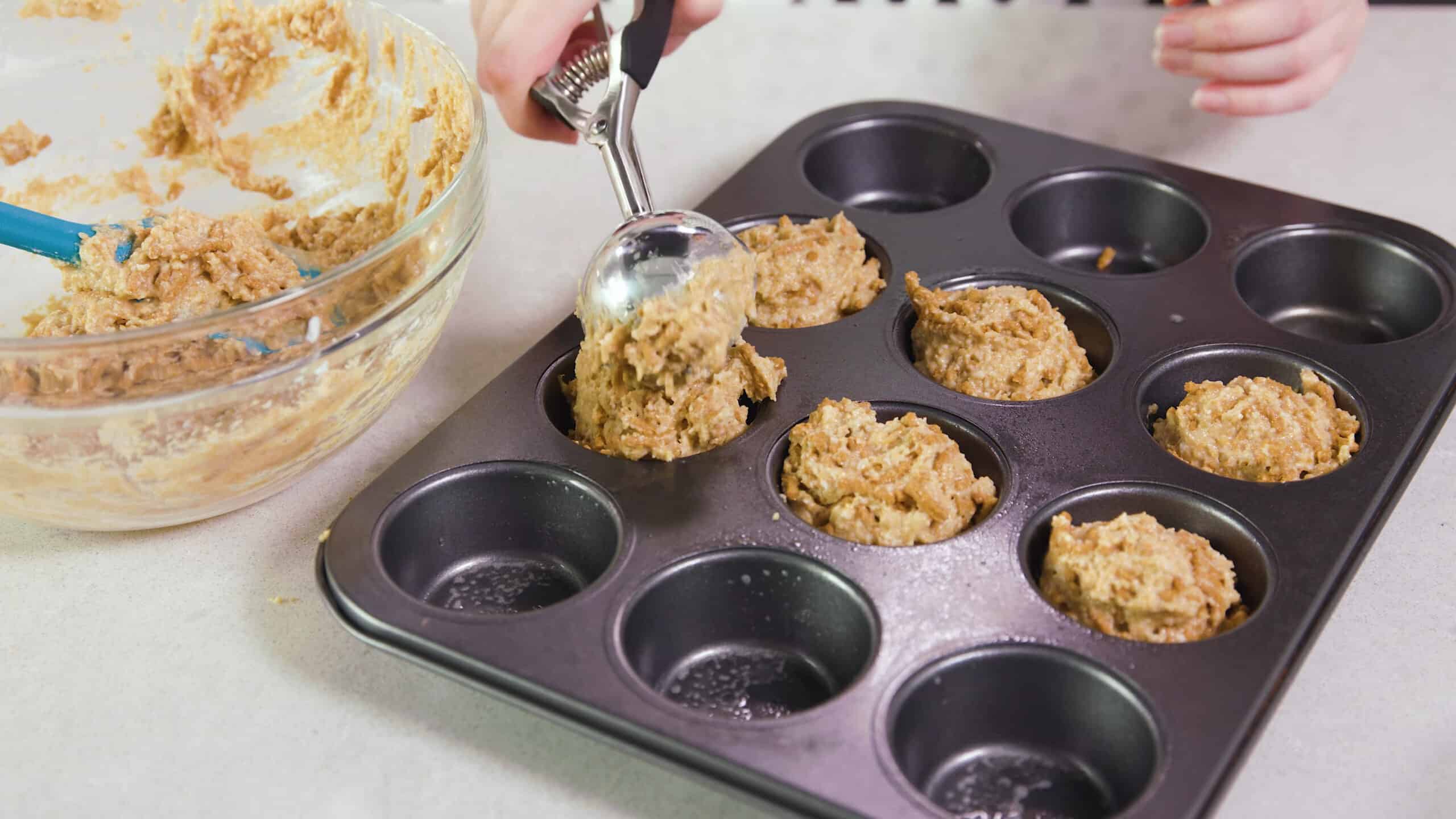 Angled view of greased metal muffin tin being filled with scoops of bran muffin mix using a metal scooper.