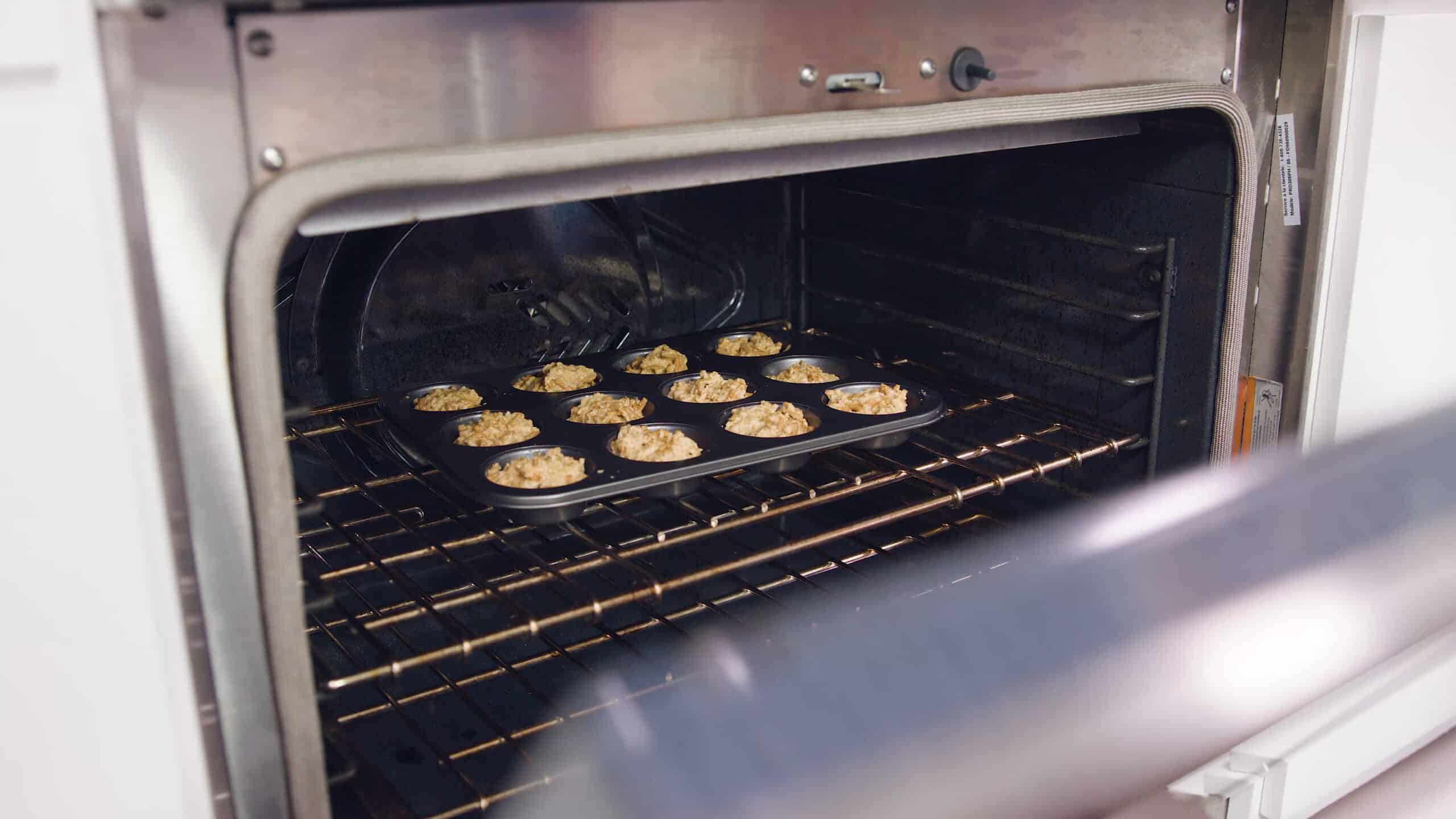Angled view of an open oven with a greased metal muffin tin filled with one dozen spots of muffin mix on a metal rack in the middle of the oven.