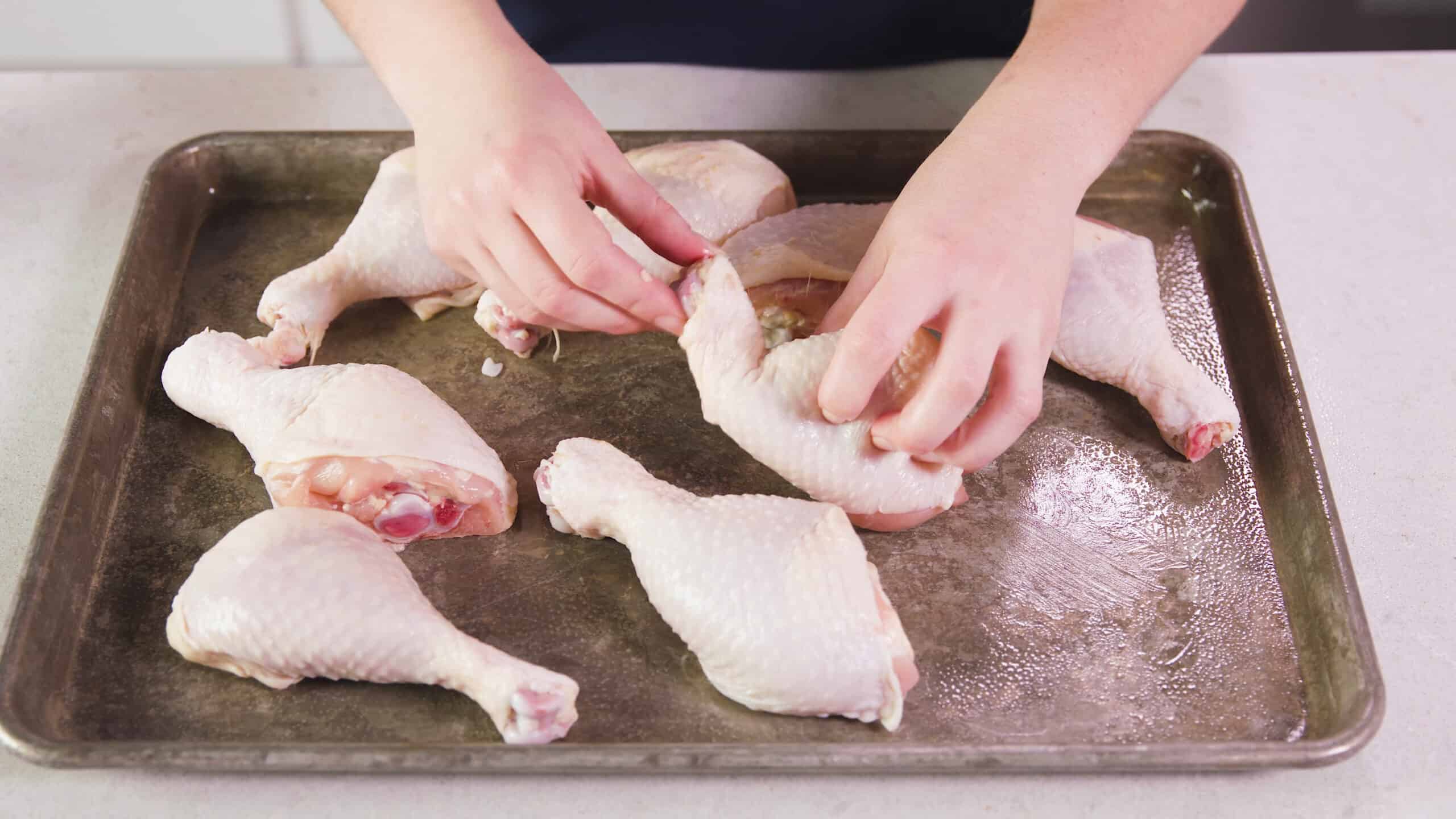 Angled view of greased metal baking sheet with eight raw chicken drumsticks placed on top one of which is being displayed by two hands to show the broken bone which is not ideal.