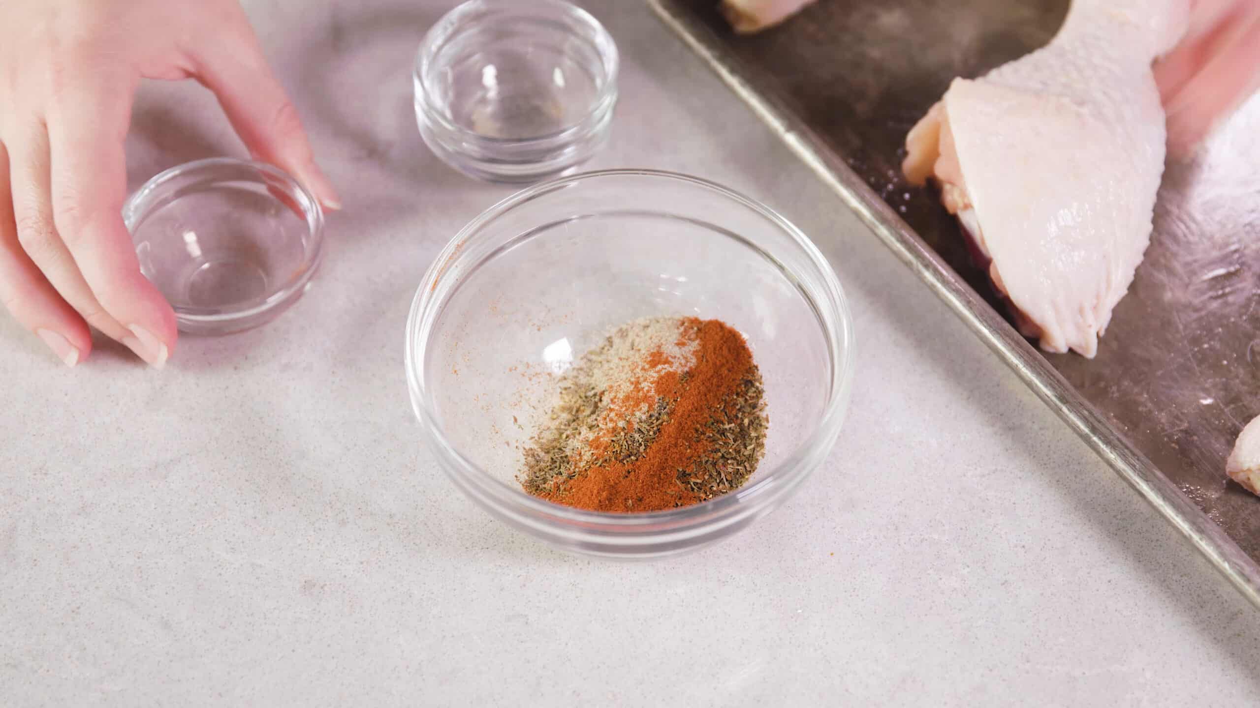 Angled view of a clear glass mixing bowl filled with dry cajun spices mixed together ready to add to the chicken which is at the ready on a metal baking sheet to the right.