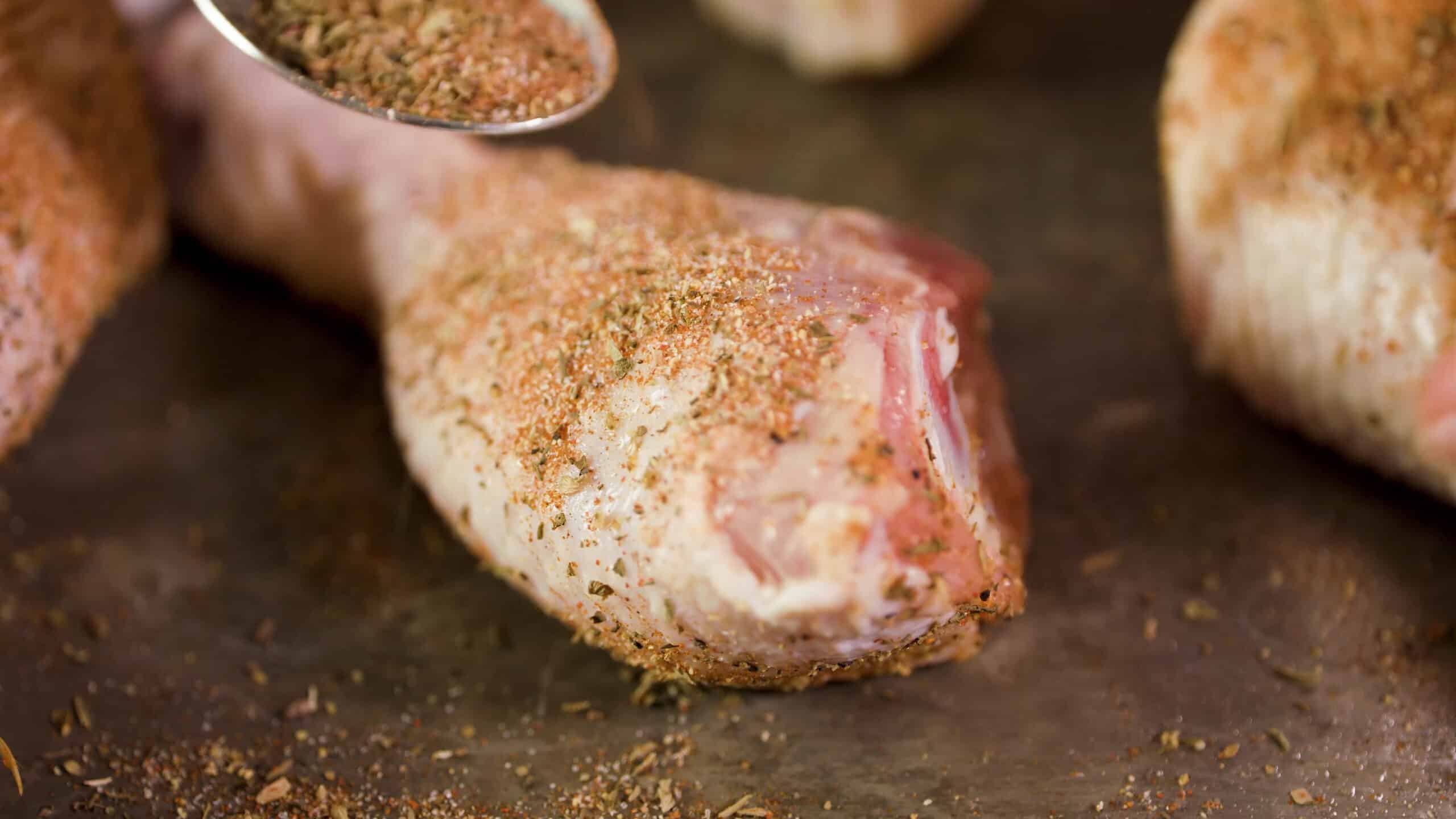 Close-up view of a single drumstick covered in dry cajun spices sprinkled from a metal spoon.