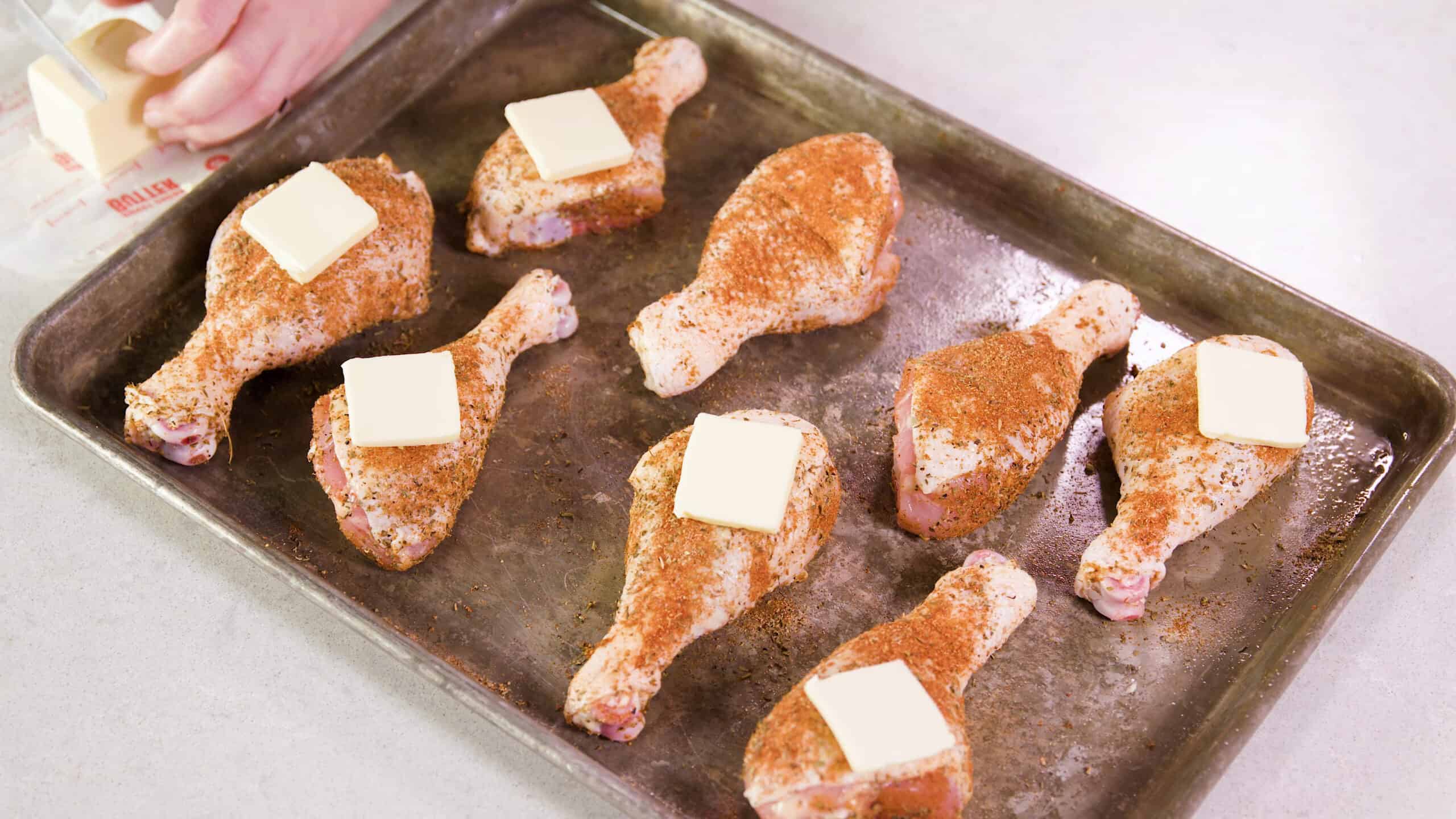 Angled view of greased metal baking sheet filled with eight raw chicken drumsticks coated with dry cajun spices and each drumstick topped with a pad of butter.
