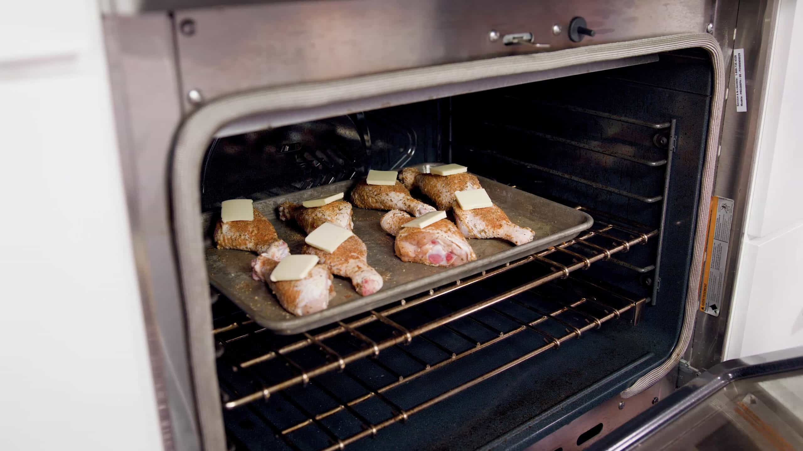 Angled view of an open oven with a greased metal baking sheet filled with eight raw chicken drumsticks coated with dry cajun spices and each drumstick topped with a pad of butter, placed on a metal rack in the middle of the oven.
