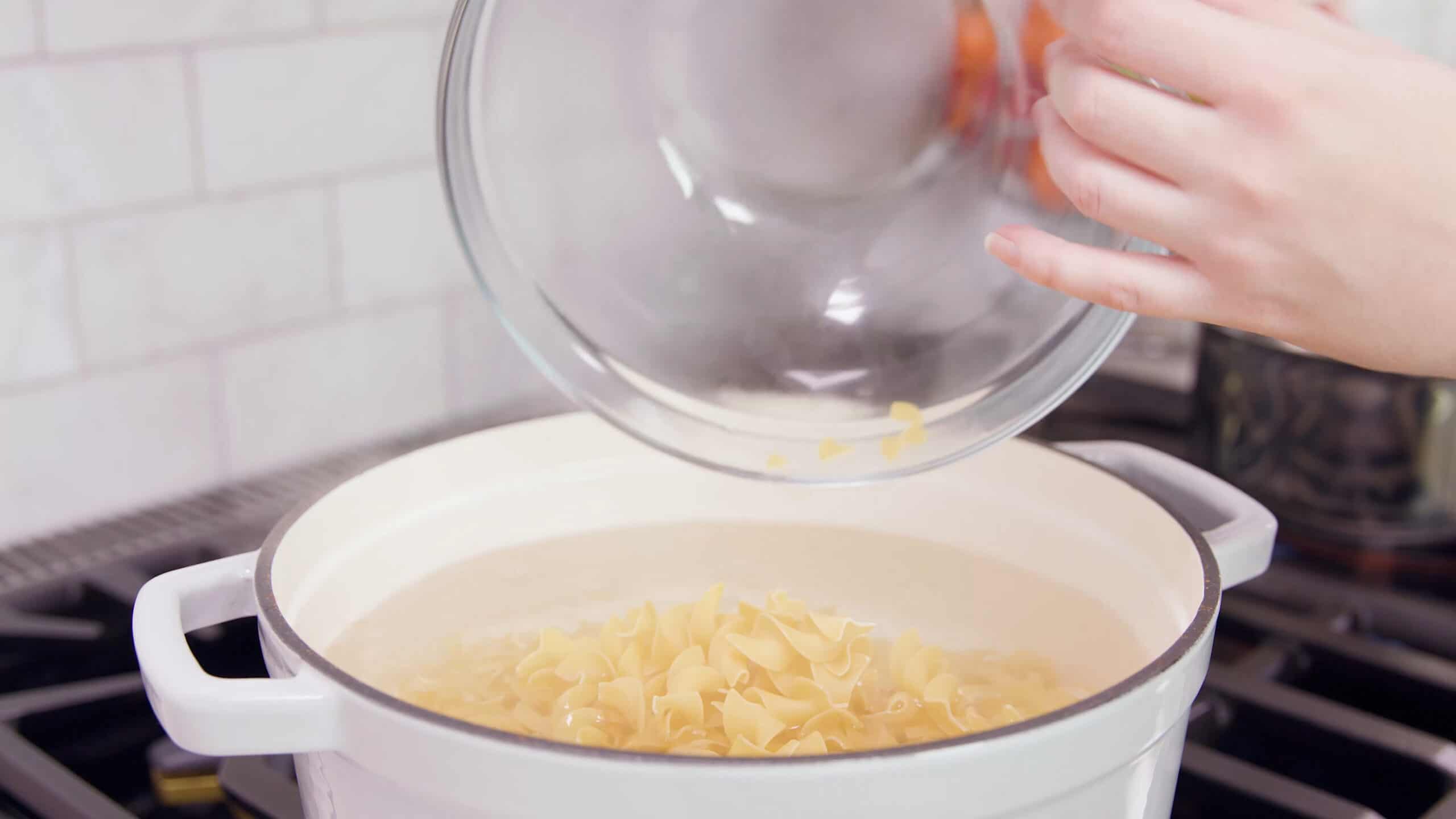 Angled view of white enamel coated cast iron pot filled with boiling water and extra wide egg noodles poured from a large clear glass mixing bowl all over a stovetop burner.