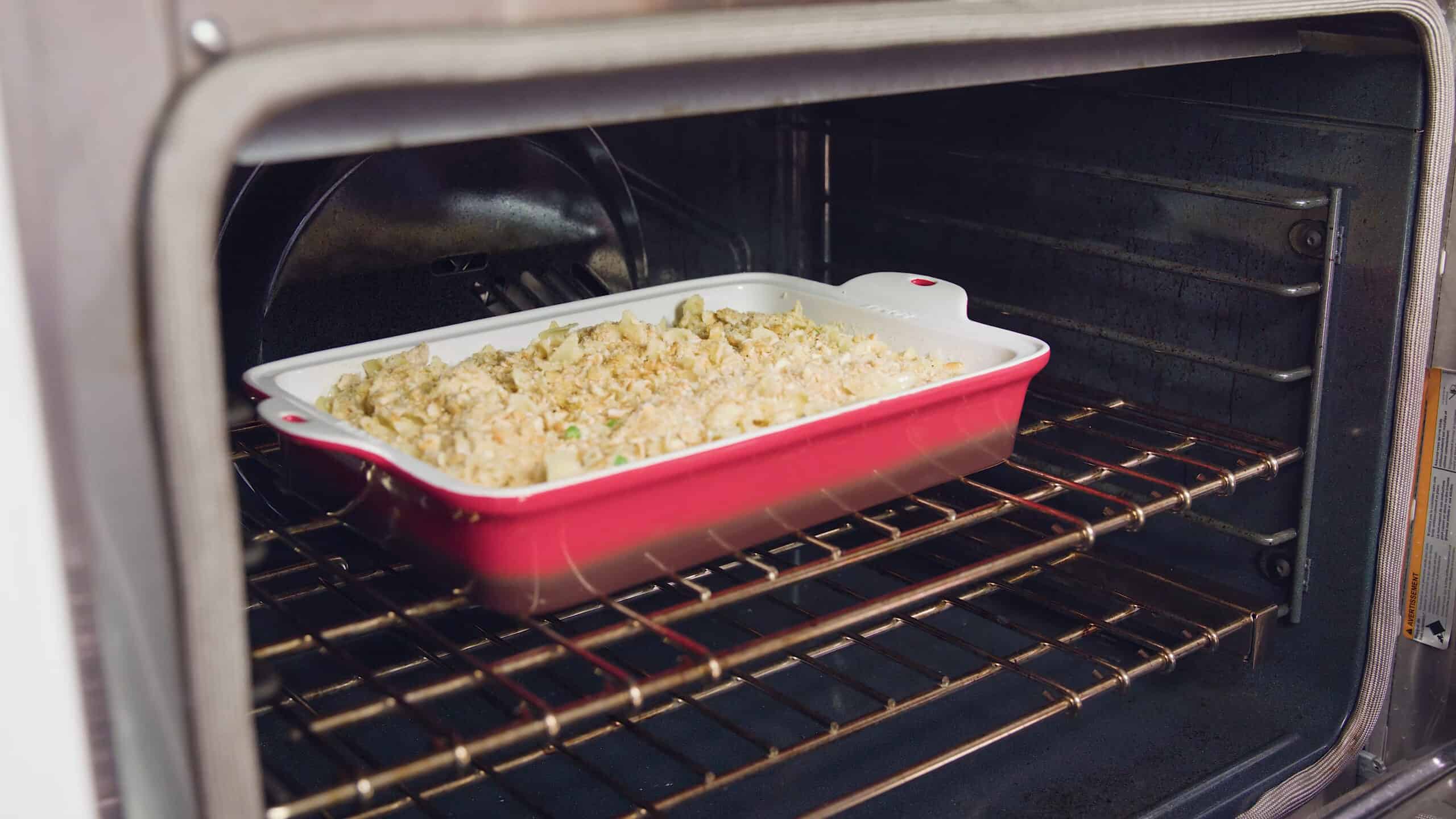 Angled view of an open oven with an enamel coated cast iron casserole dish filled with creamy chicken casserole mixture, egg noodles, and golden cracker topping all on a metal rack in the middle of the oven.