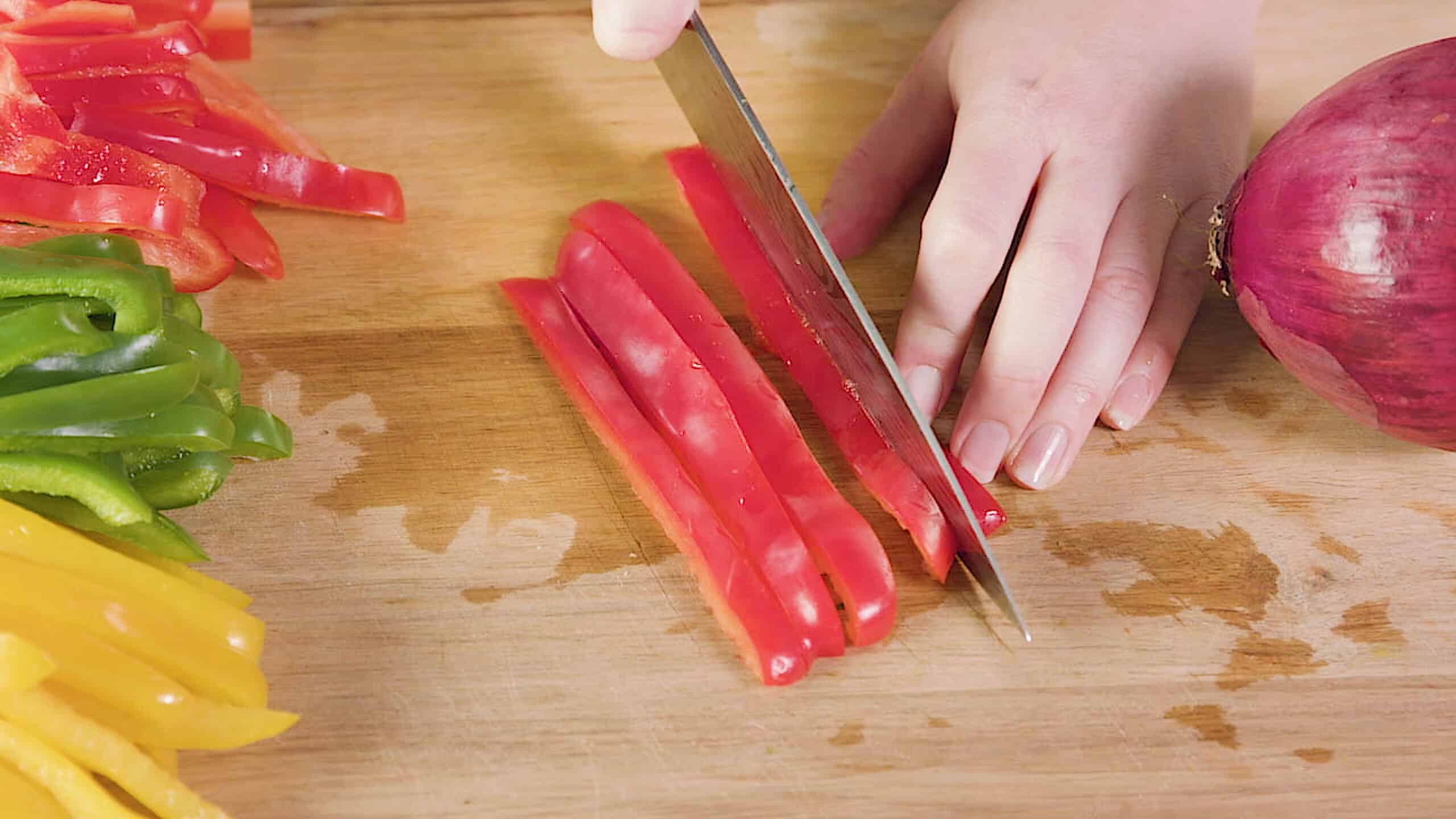 Close-up view of a wooden cutting board with red peppers being sliced by hand with a metal kitchen knife framed by sliced red, yellow, and green peppers and a whole red onion.
