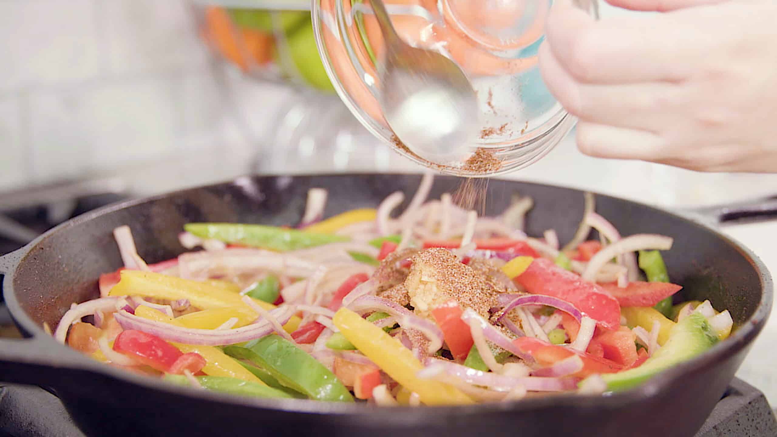 Close-up view of a cast iron skillet filled with sliced red onion and sliced red, yellow, and green peppers topped with minced garlic and various spices poured from a small clear glass mixing bowl scooped out using a metal spoon.