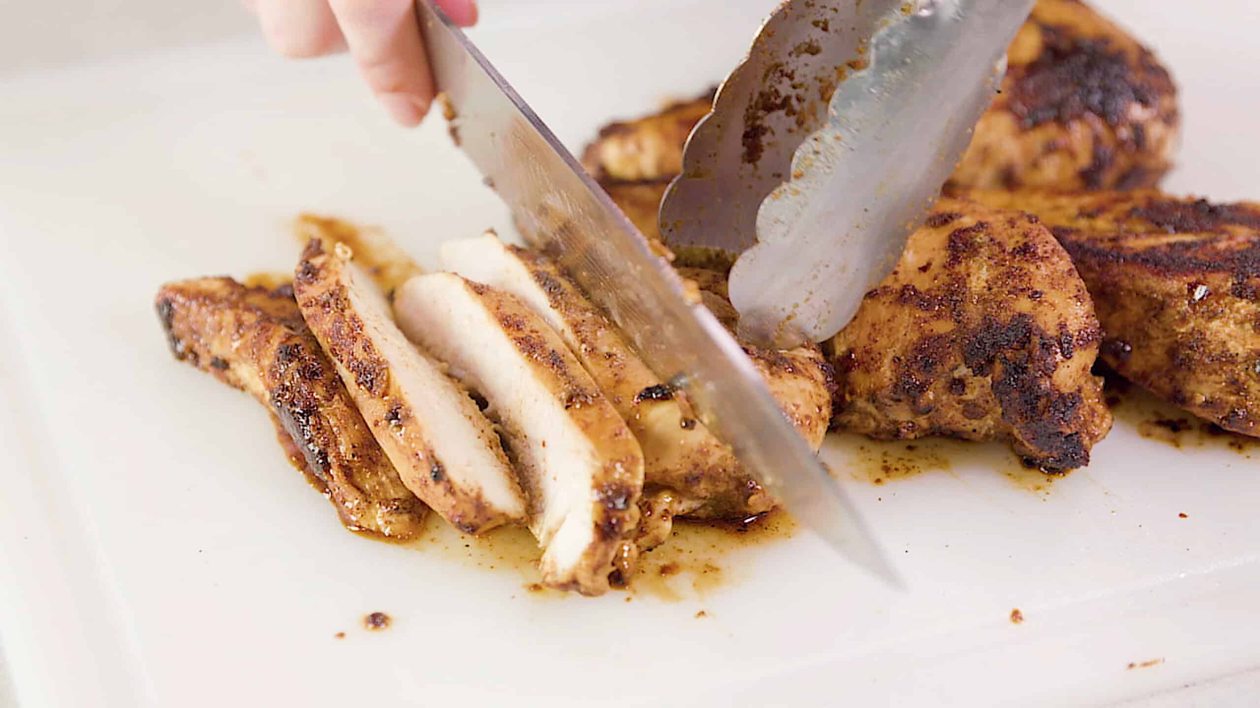 Close-up view of a white plastic cutting board topped with cooked seasoned chicken breasts being sliced by hand using a metal kitchen knife.