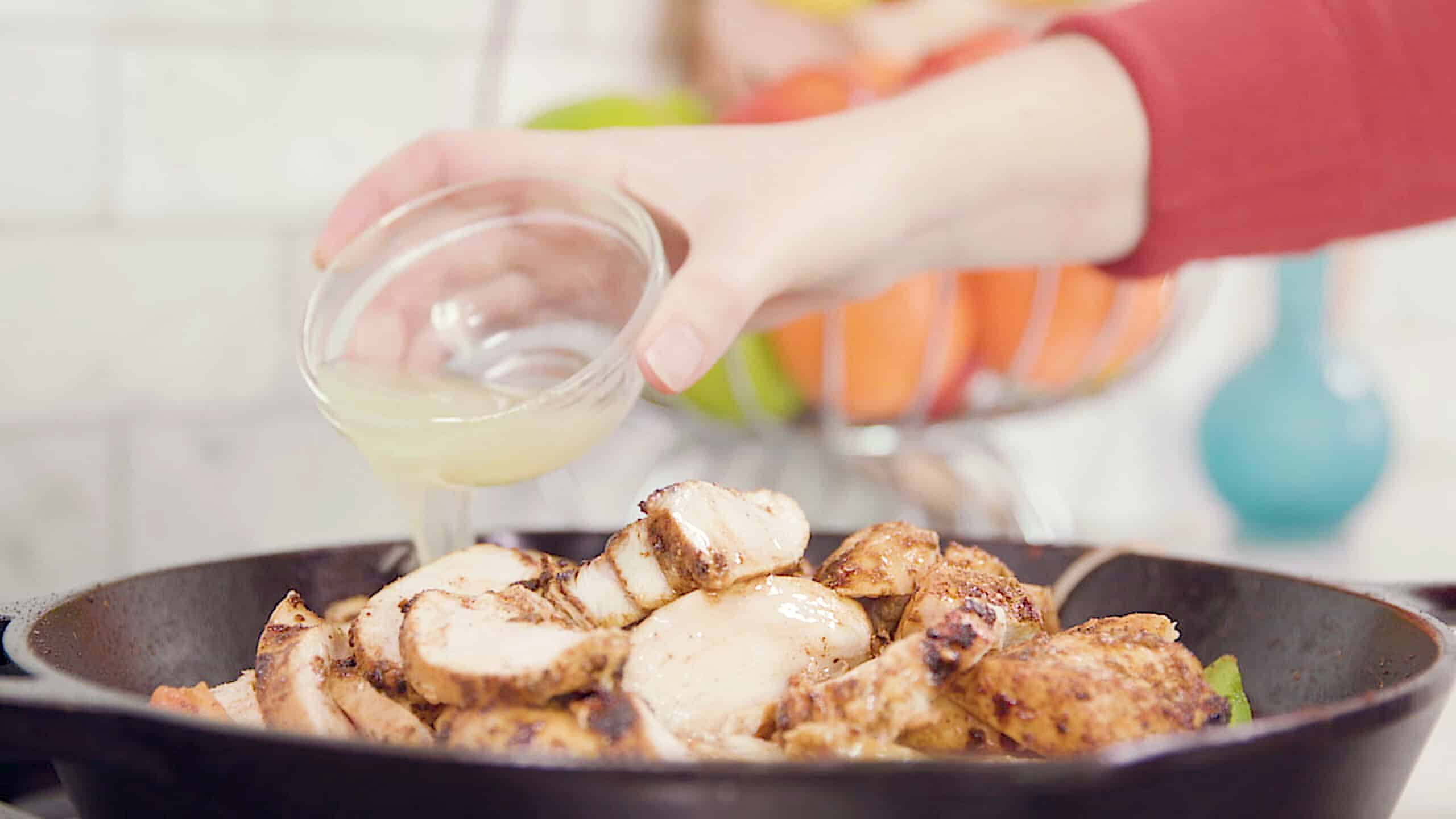 Side view of cast iron skillet filled with sliced seasoned chicken breast and topped with lime juice poured by hand from a small clear glass mixing bowl.