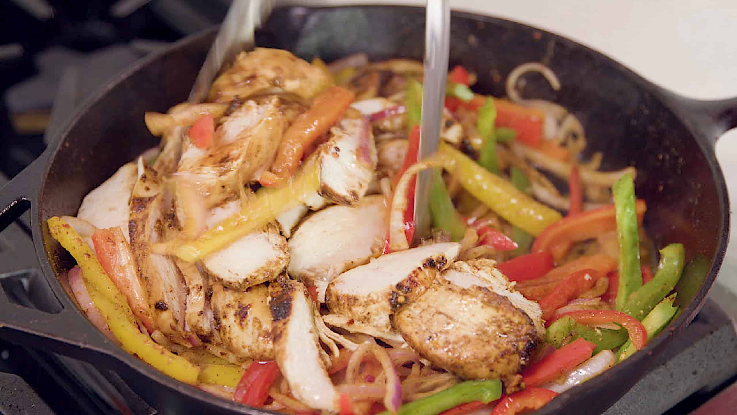 Overhead view of cast iron skillet filled with sliced seasoned chicken breast and sliced red onion and sliced red, yellow, and green peppers tossed using metal tongs to combine.