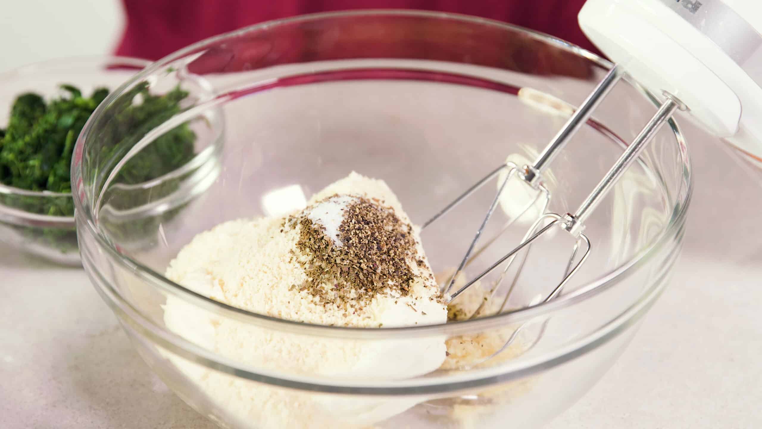 Angled view of a large clear glass mixing bowl filled with ingredients for the artichoke dip including cream cheese, salt and pepper, parmesan cheese and minced garlic with wire beaters in a hand mixer at the ready to combine.